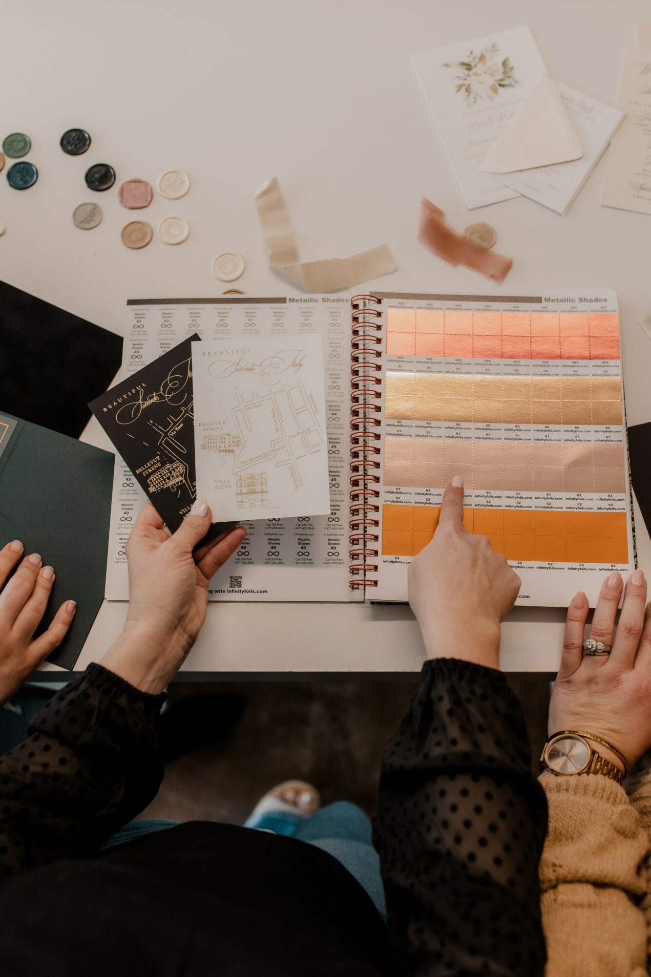 Person pointing at color swatches in a catalog, with paint samples and paper on a white table.