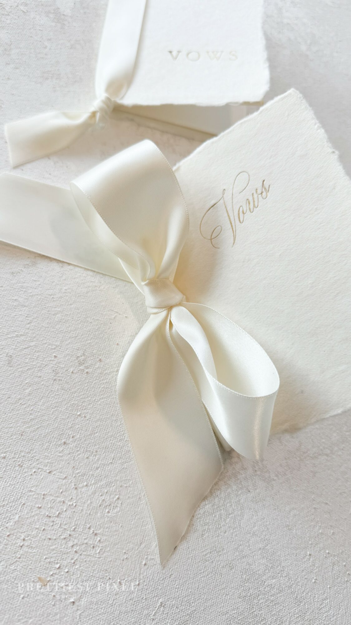 Cream-colored gift box with a satin ribbon bow, textured paper, and elegant script text, on a light surface.