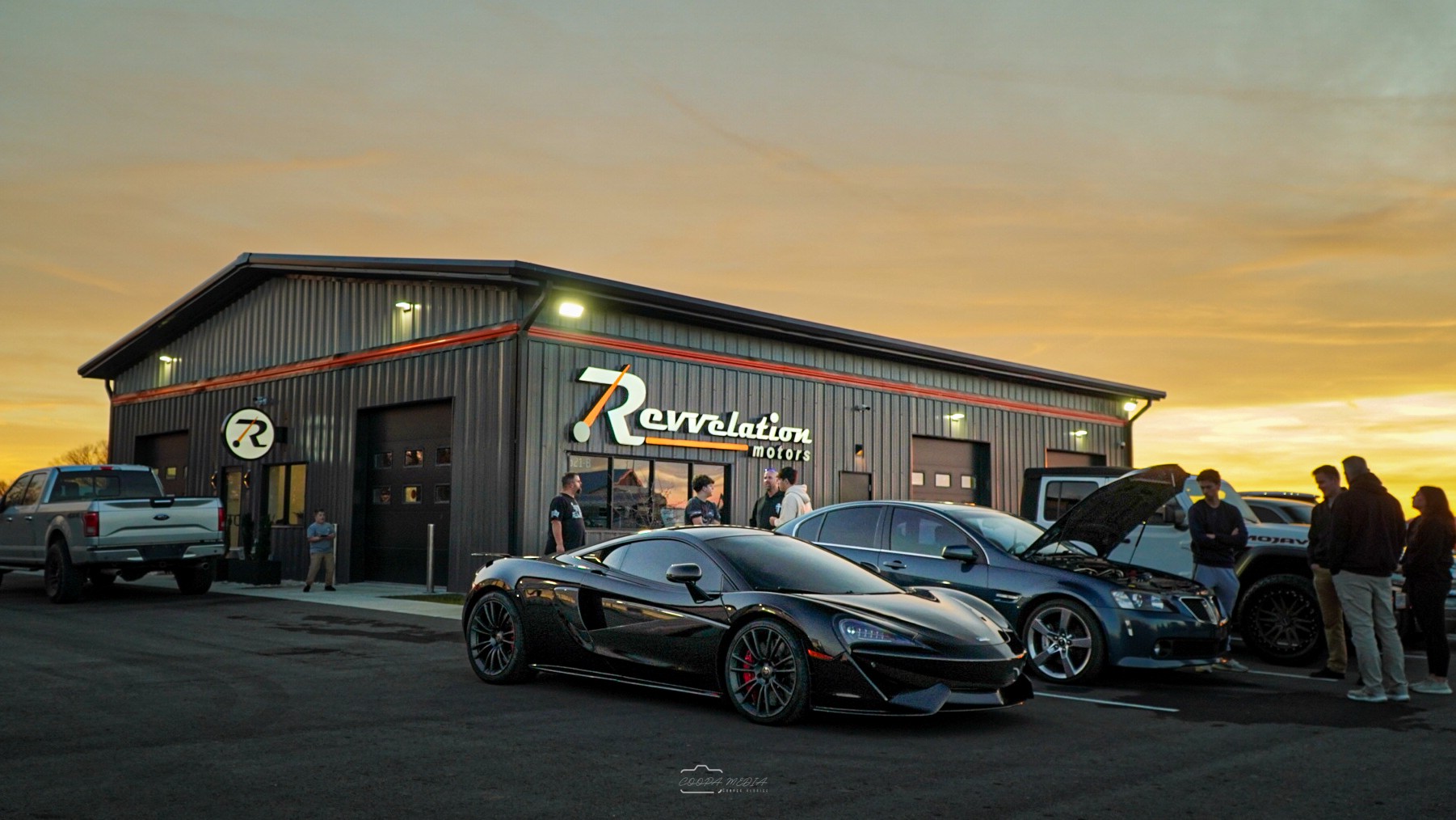 A building with a sign reading 'Revelation' at sunset, cars parked outside, people gathered around, some inspecting vehicles.