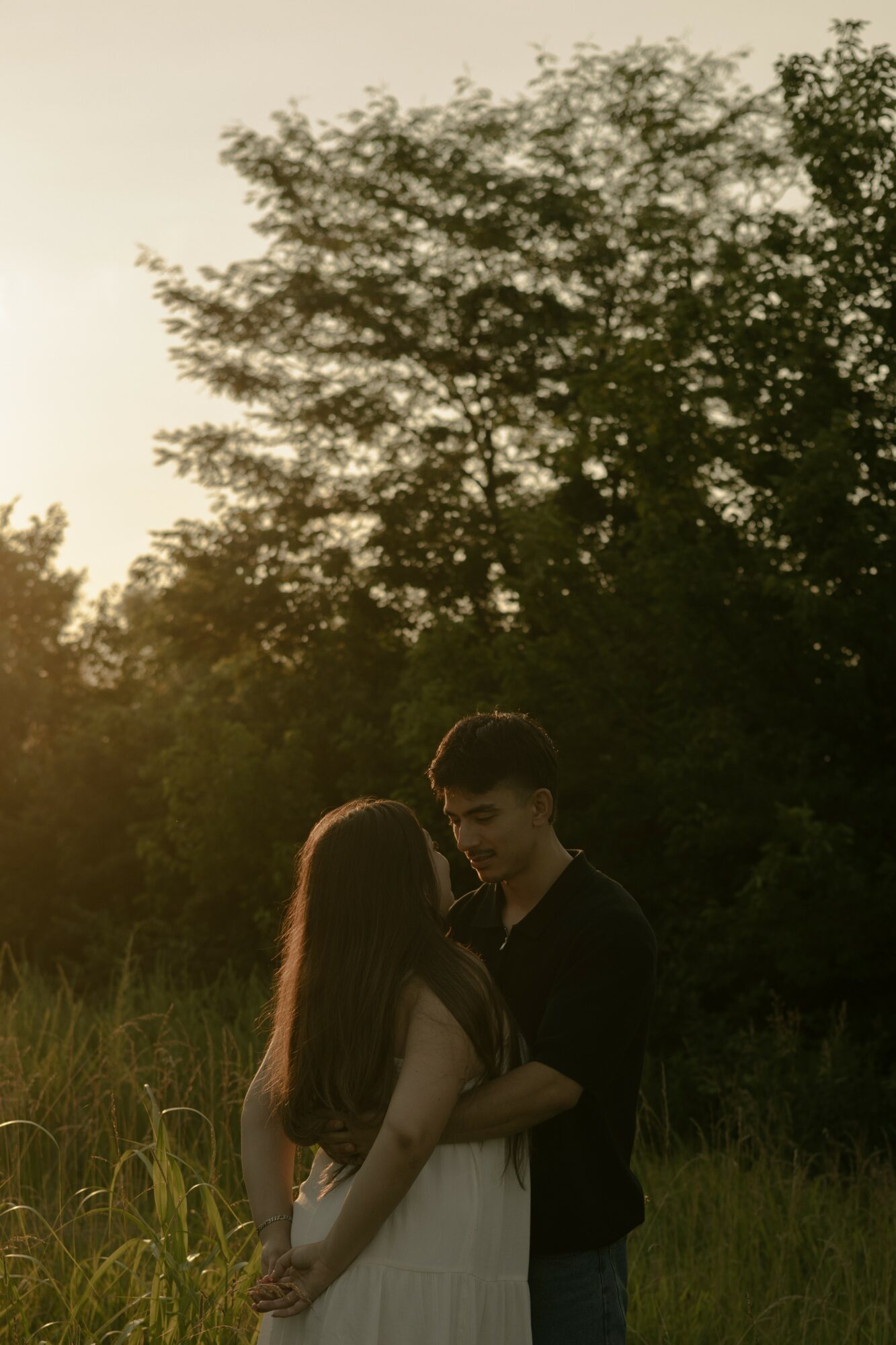 A young man and woman stand close together outdoors, with trees and grass in the background during sunset.