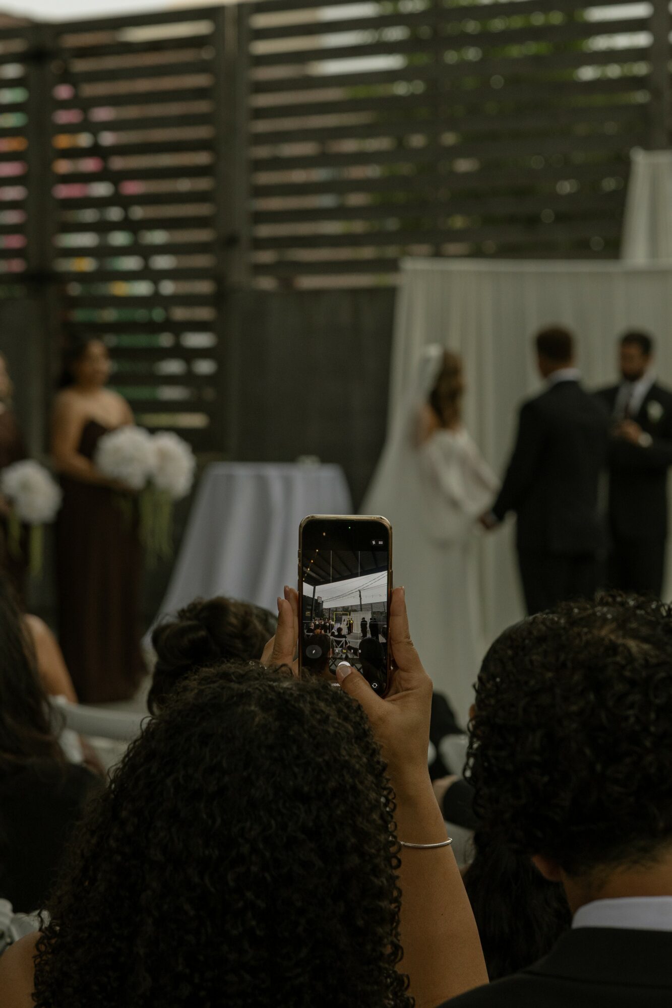 Person taking photo of wedding ceremony with smartphone, guests in foreground, bride and groom at altar, outdoor setting.