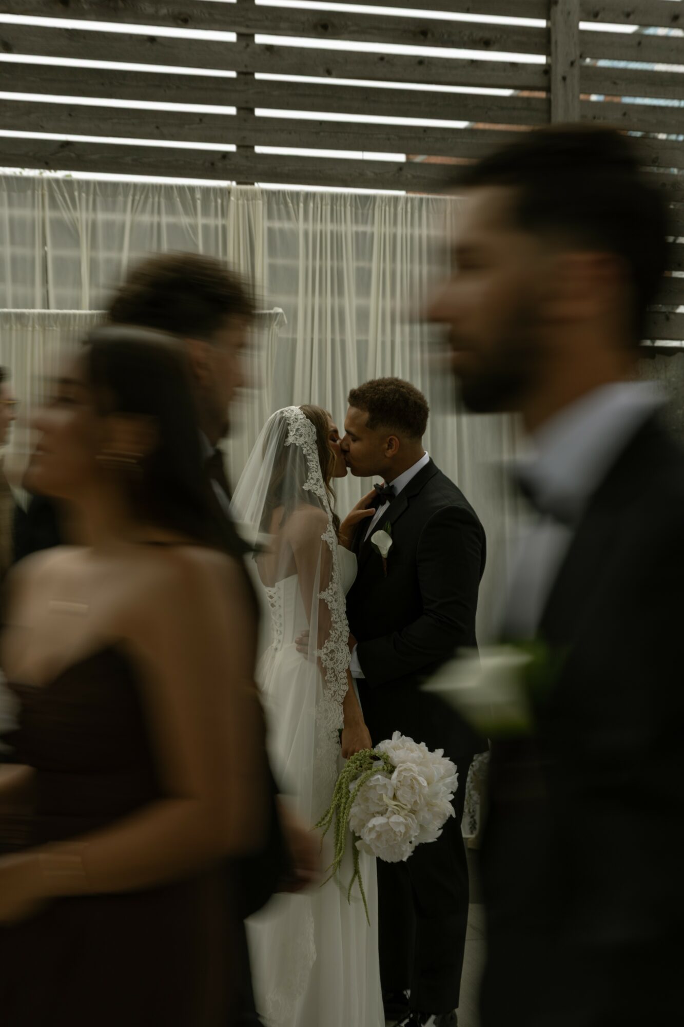 Bride and groom face each other during wedding ceremony, surrounded by blurred guests, with a wooden fence and curtains in background.