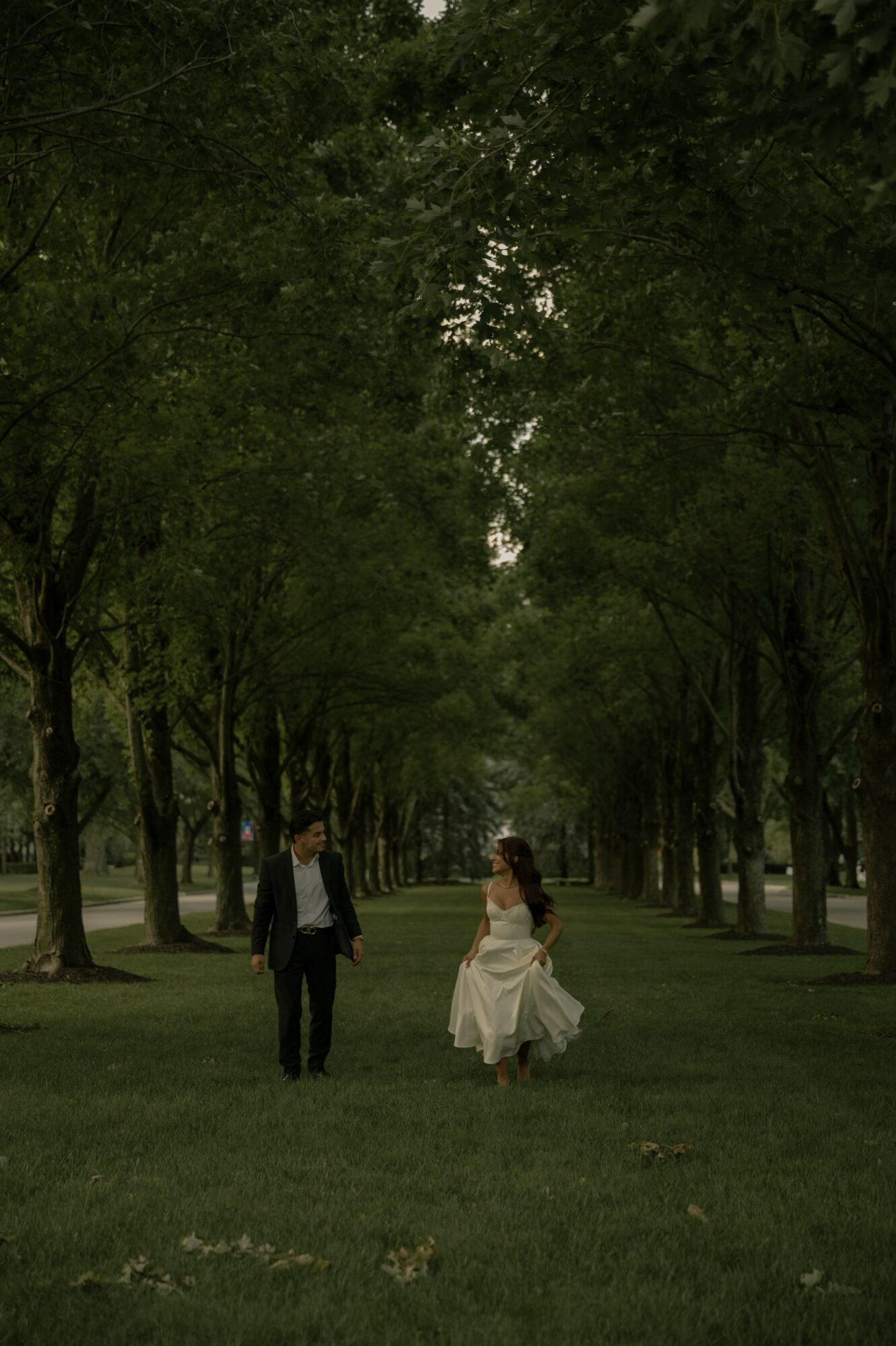 Two people walking in a park with tall trees and green grass, one in a suit and the other in a white dress.