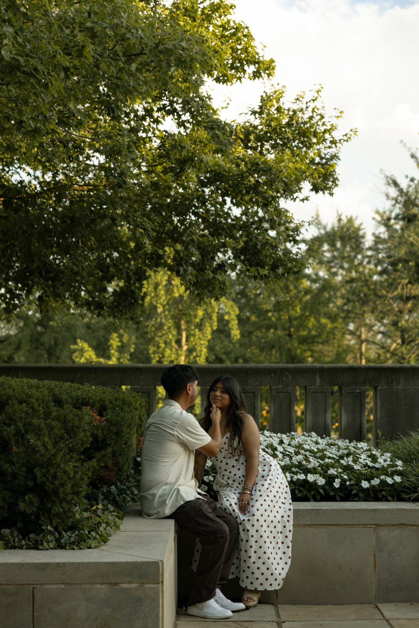 A man and woman sit on a low wall outdoors, surrounded by greenery and white flowers, engaging in conversation.