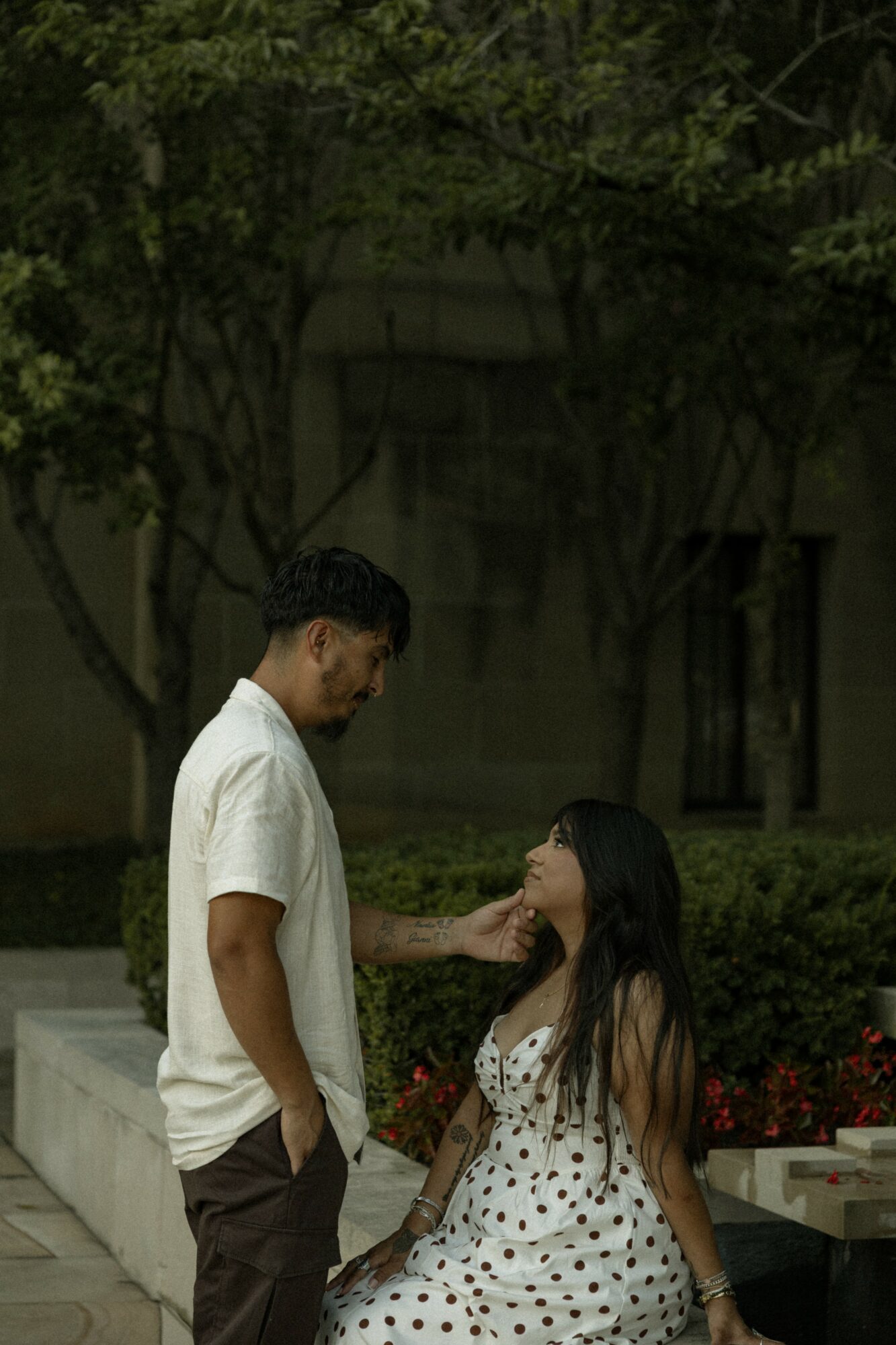 A man and woman outdoors, the man touching the woman's face, surrounded by trees and bushes.