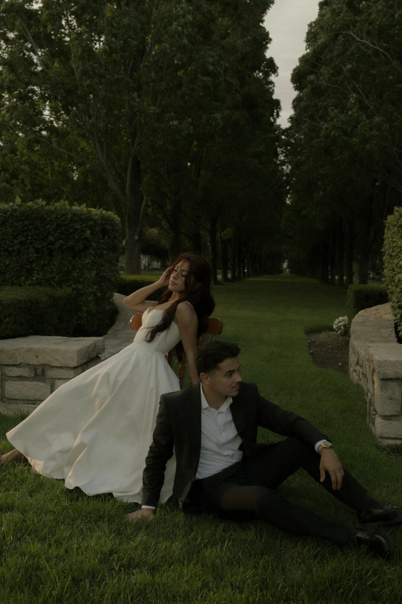 A couple in formal attire sitting on grass in a park with tall trees and hedges, woman in a white dress, man in a black suit.