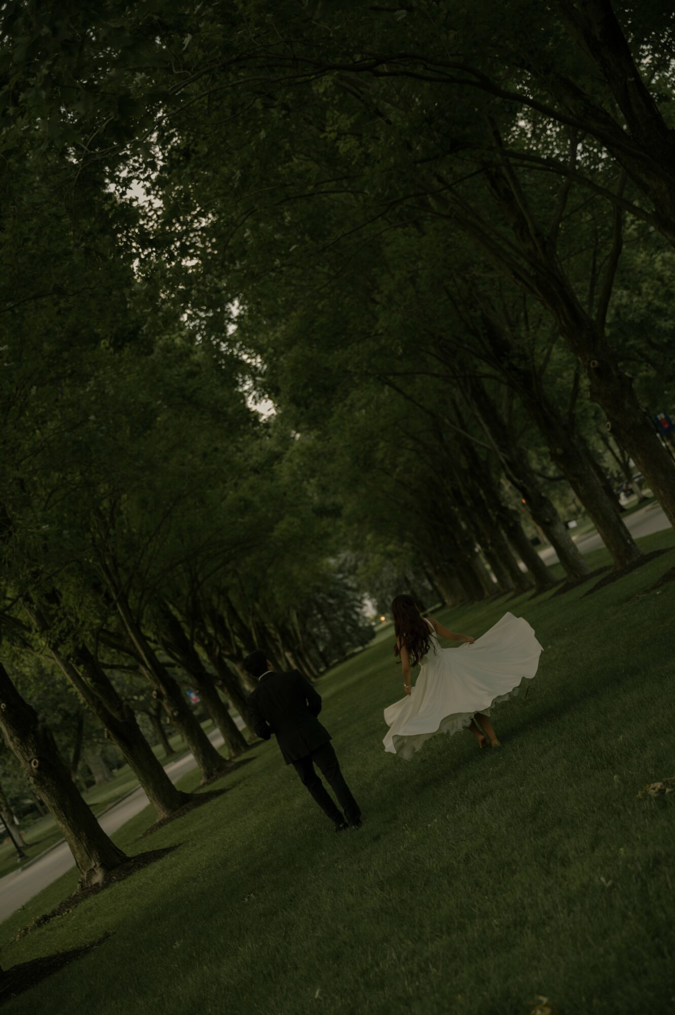A person in a dark outfit and a person in a white dress walk through a park with tall trees and green grass.