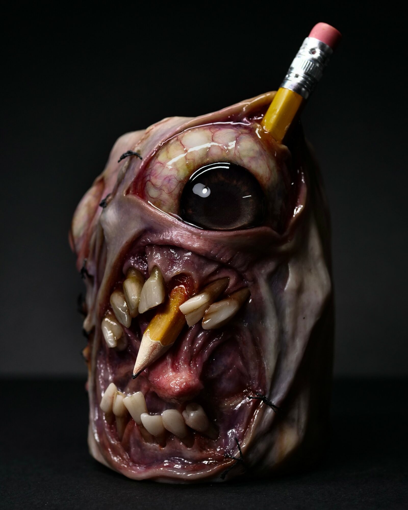 A grotesque, monster-like face with large eye, sharp teeth, and a pencil in its head, set against a dark background.