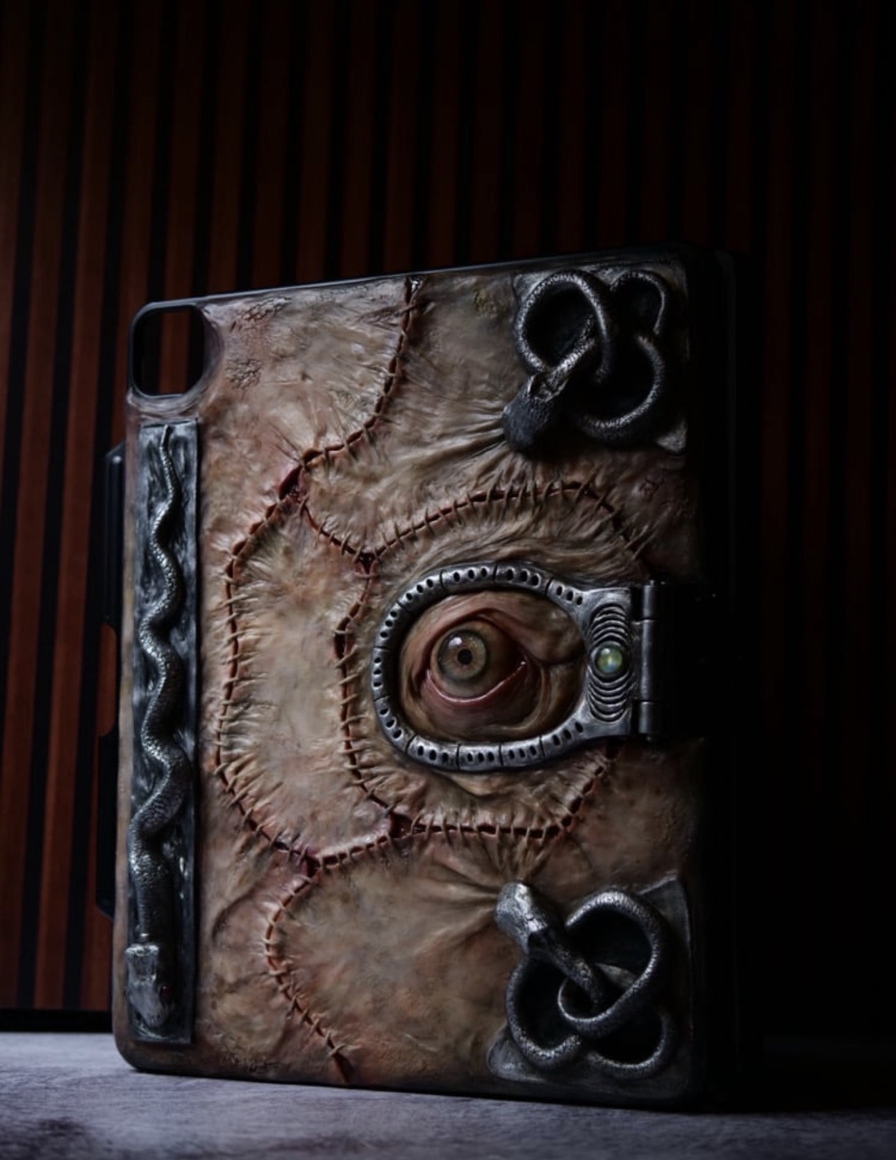 A textured, artistic object with a central eye, surrounded by metallic and leather-like elements, against a dark background.