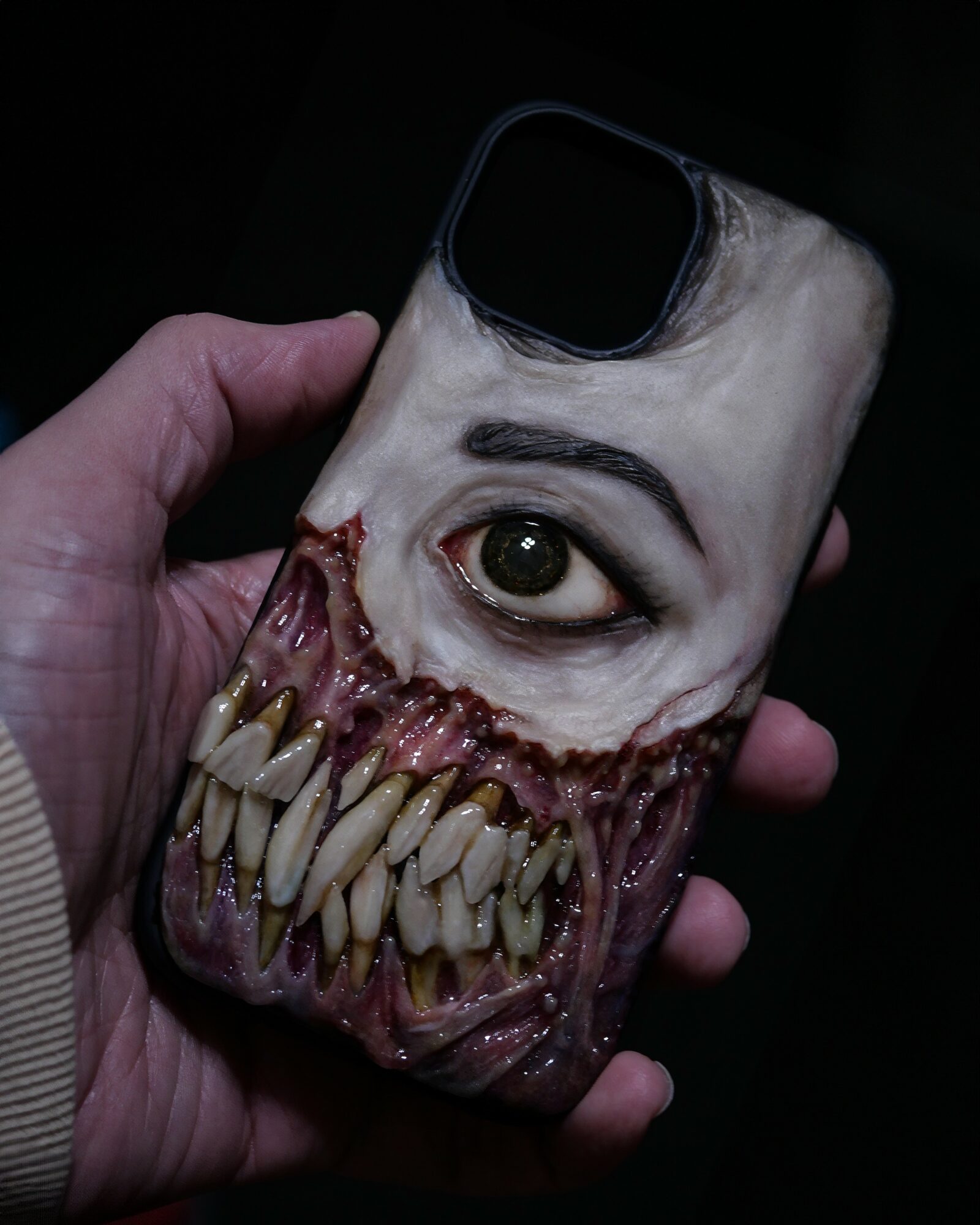 Hand holding a phone case with a horror-themed design of a distorted face with an eye and sharp teeth.