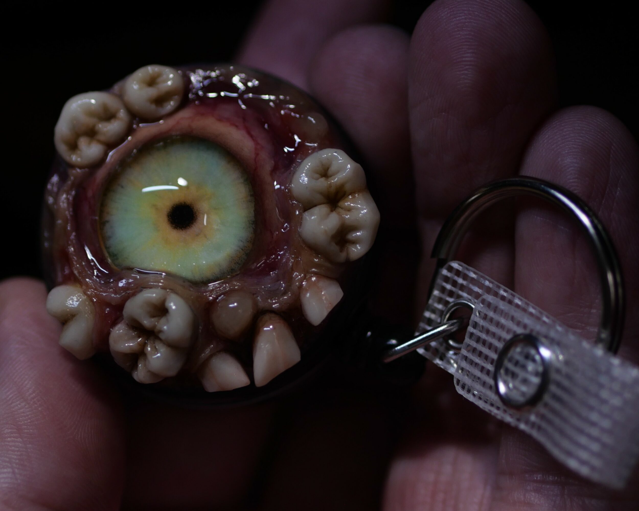 Object resembling an eye with surrounding teeth, held by a hand, attached to a keychain, dark background.