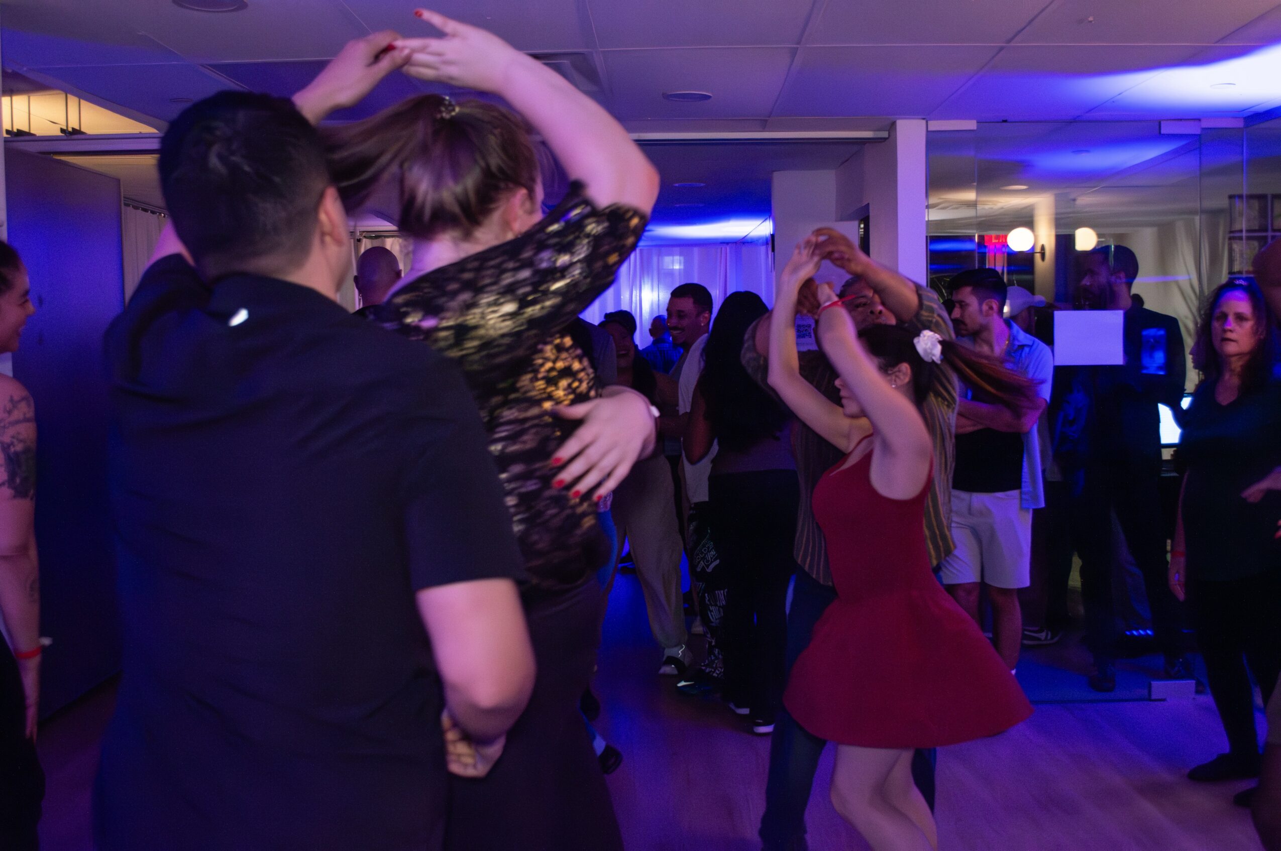 People dancing at a party with blue and purple lighting, some holding hands and embracing, in a lively indoor setting.