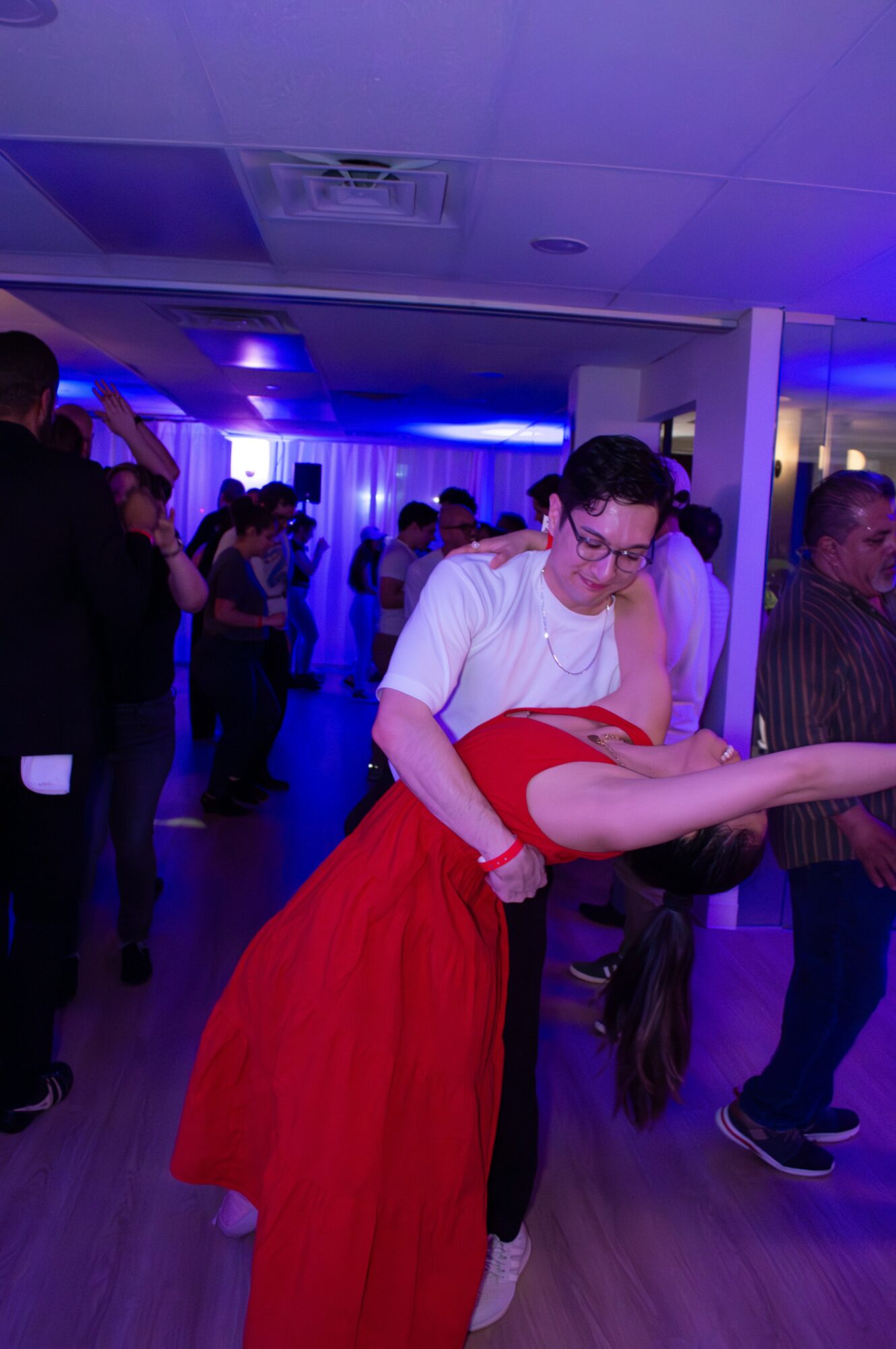 Person in white shirt dancing with a person in a red dress at a party, with other guests in the background.