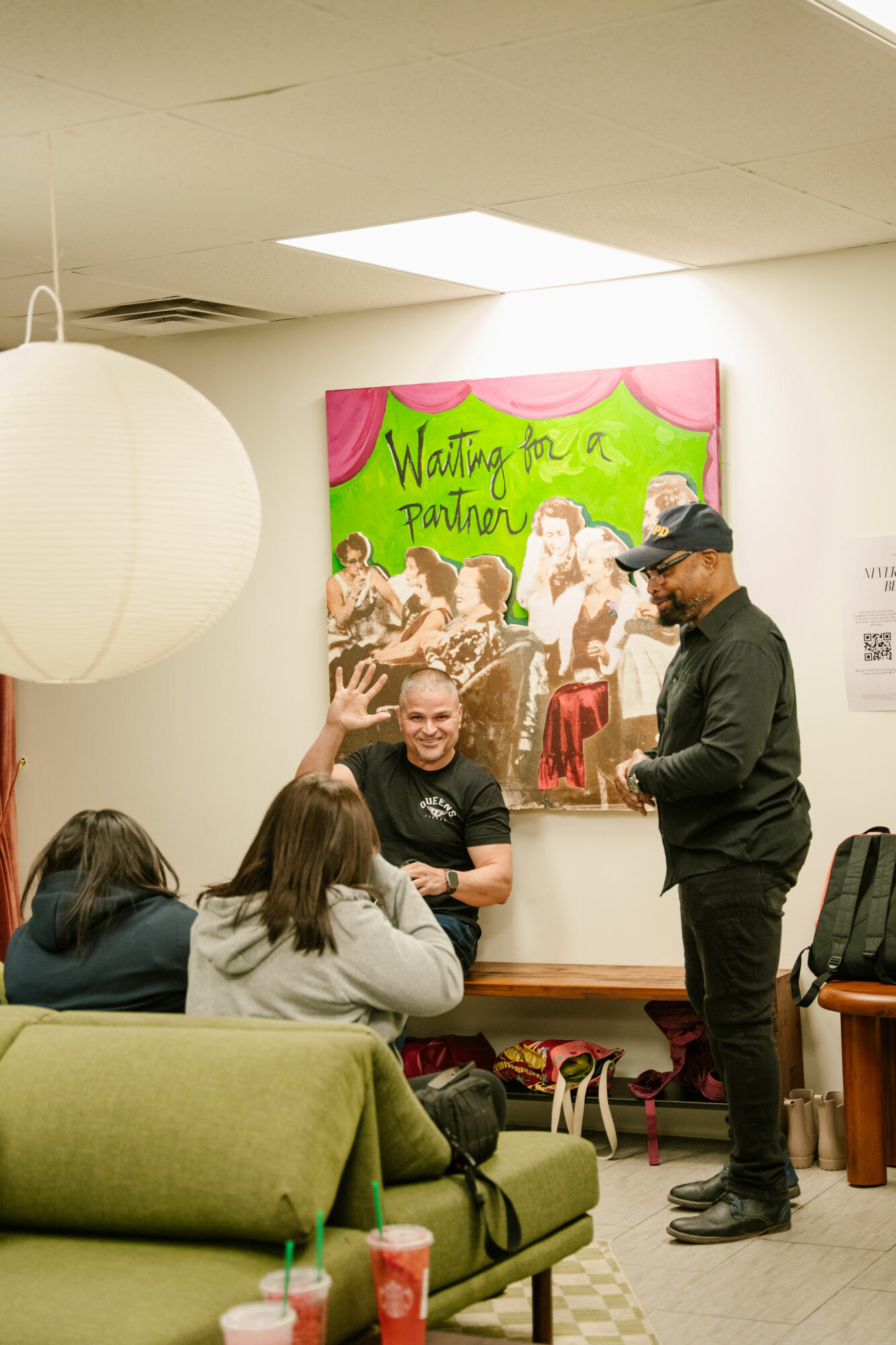 Group of people in a room with a colorful painting on the wall, some sitting and others standing, engaging in conversation.