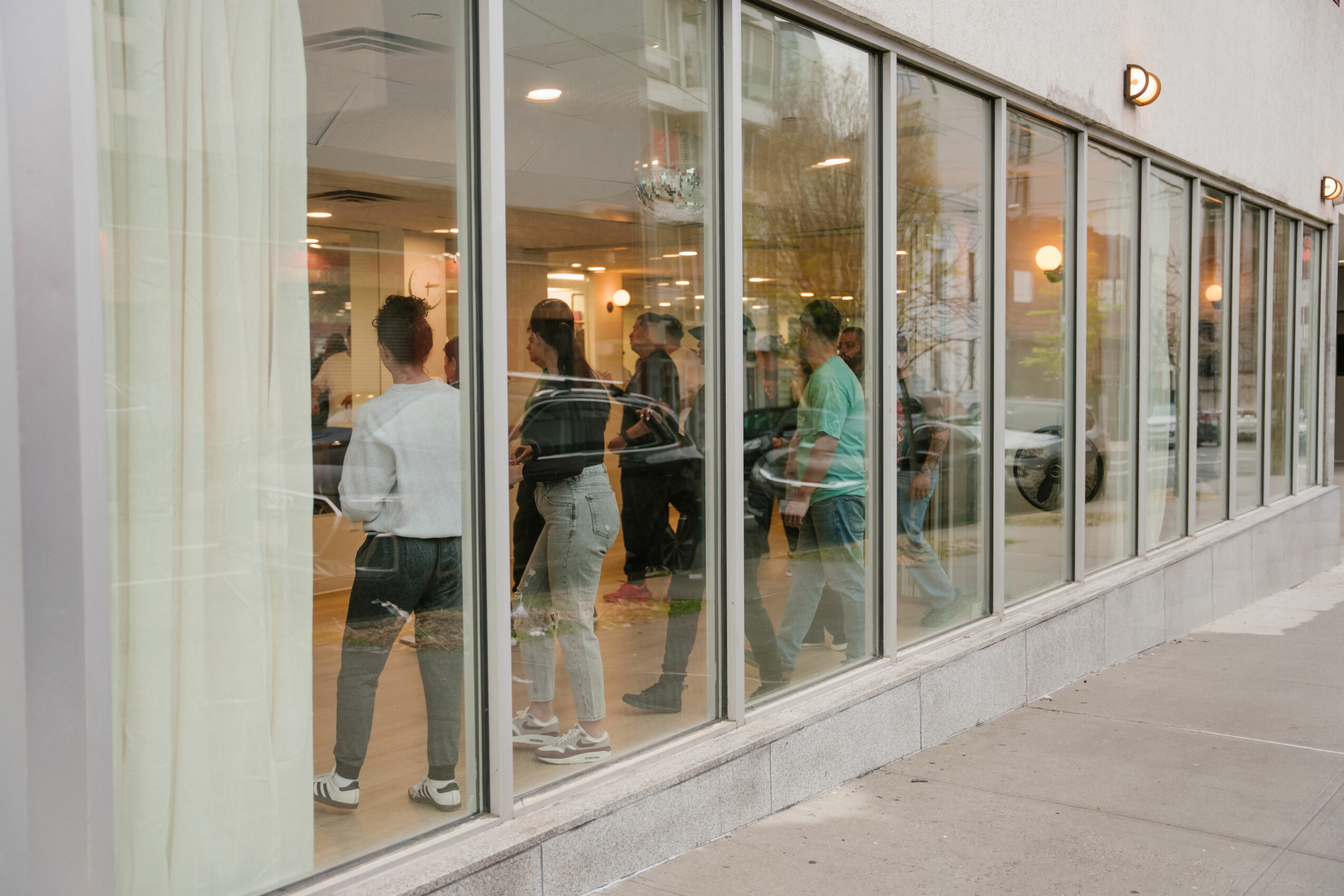 People inside a cafe or restaurant seen through large glass windows, some standing and talking, with warm lighting inside.