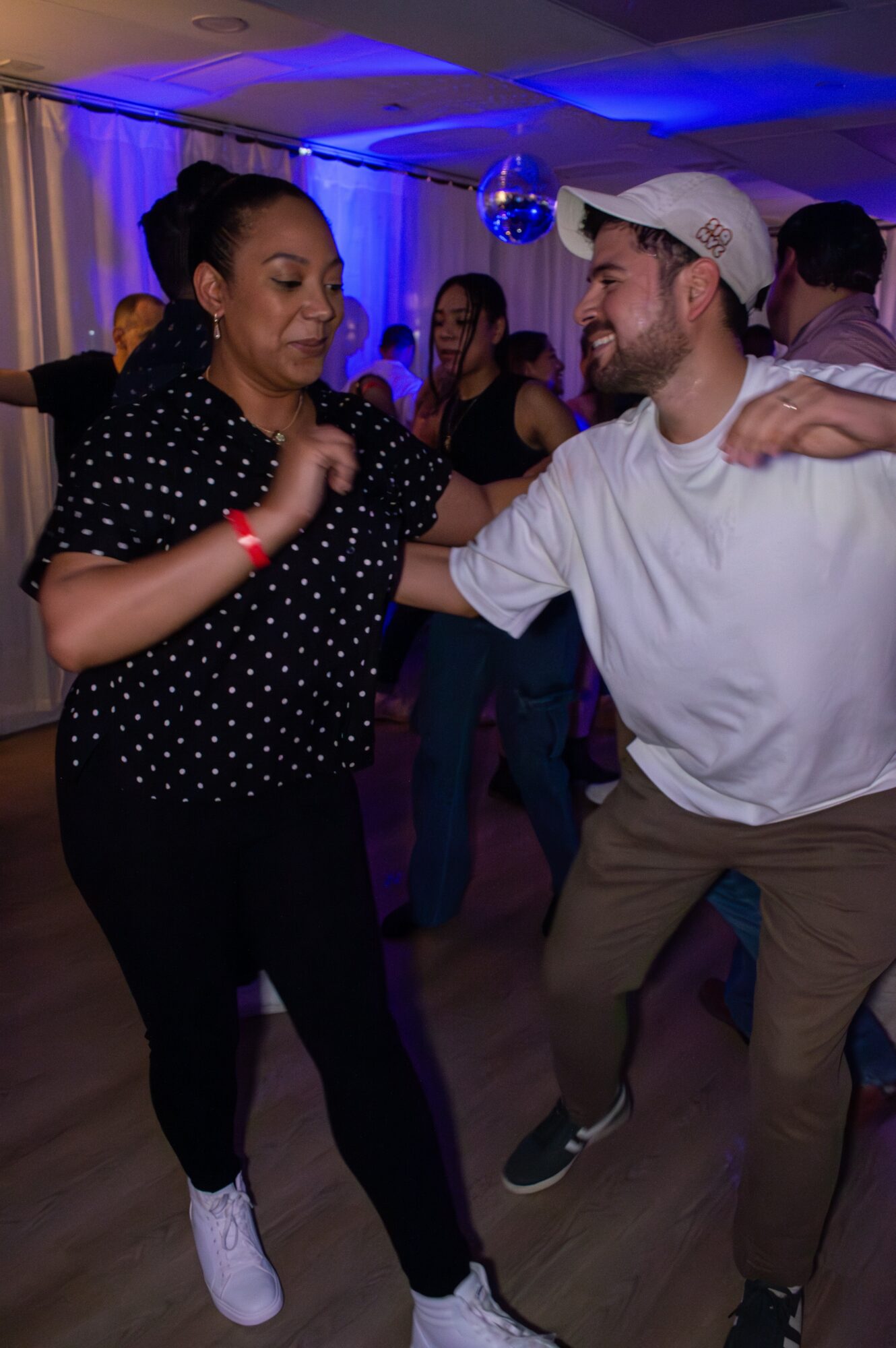 Two people dancing together at a party, smiling and holding hands, with others in the background under blue lighting.