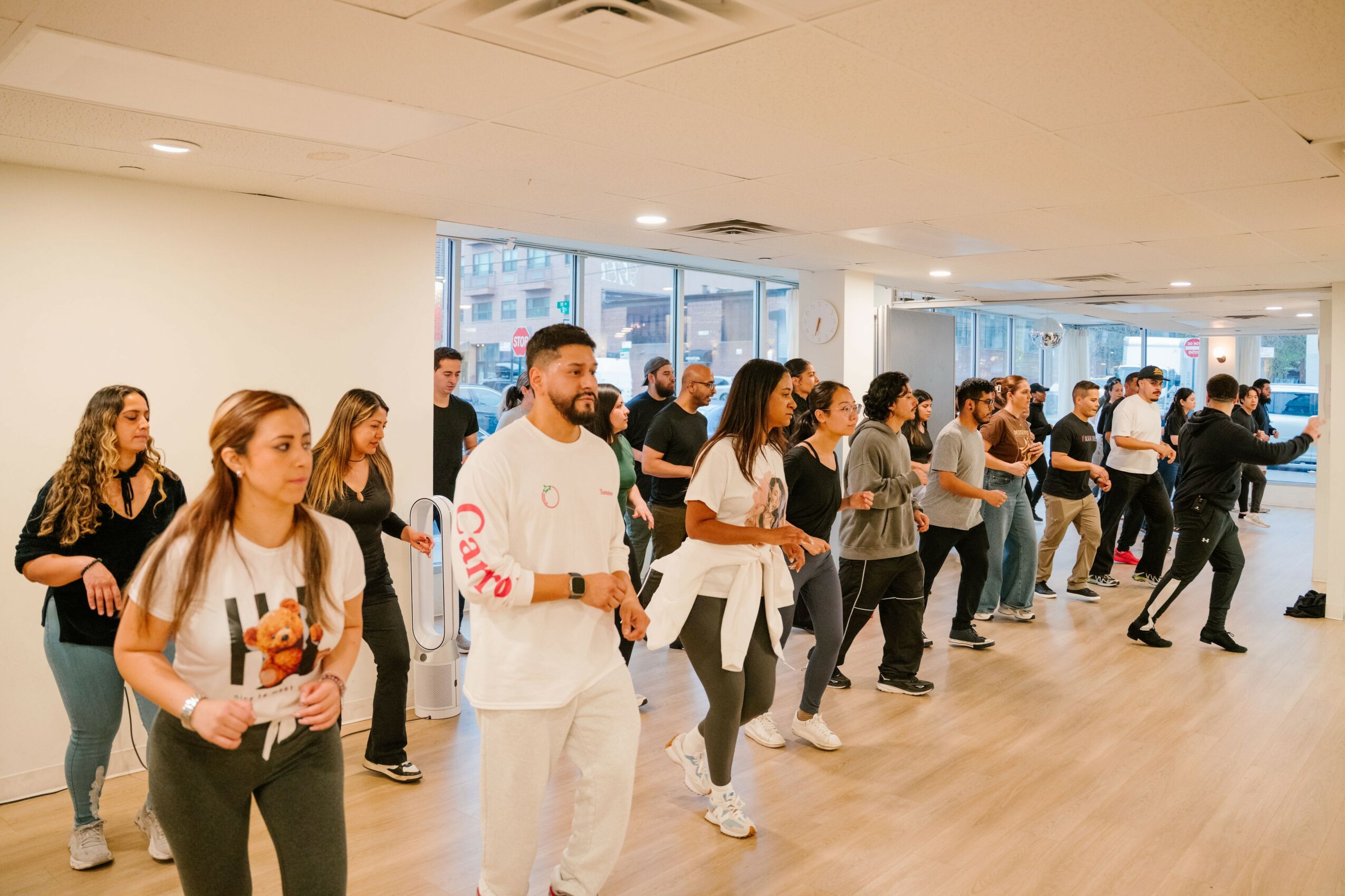 Group of people dancing in a spacious room with large windows and wooden floor, some holding drinks, lively atmosphere.