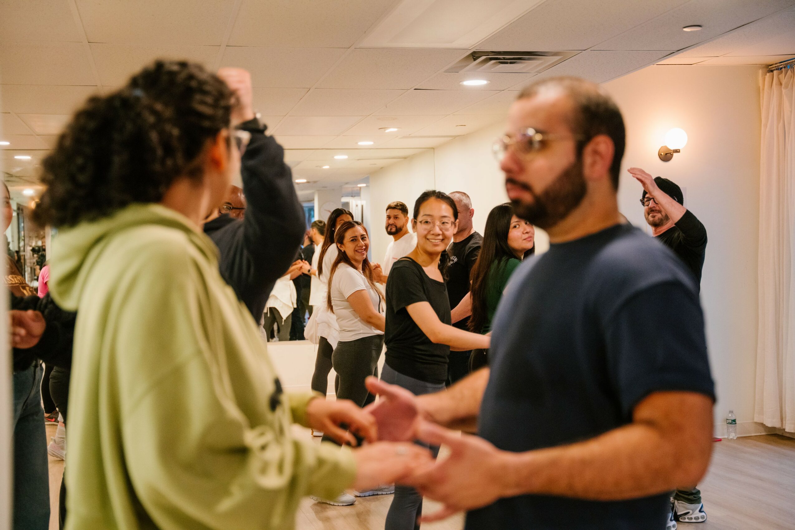Group of people dancing and socializing indoors, some holding hands, smiling, and engaging with each other.
