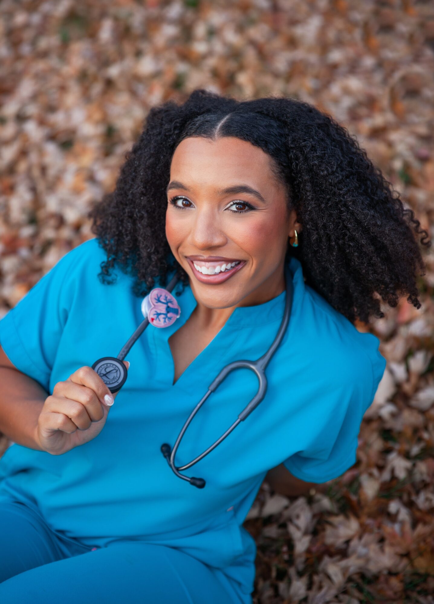 Female healthcare worker smiling outdoors, wearing blue scrubs and stethoscope, with fallen leaves in background.