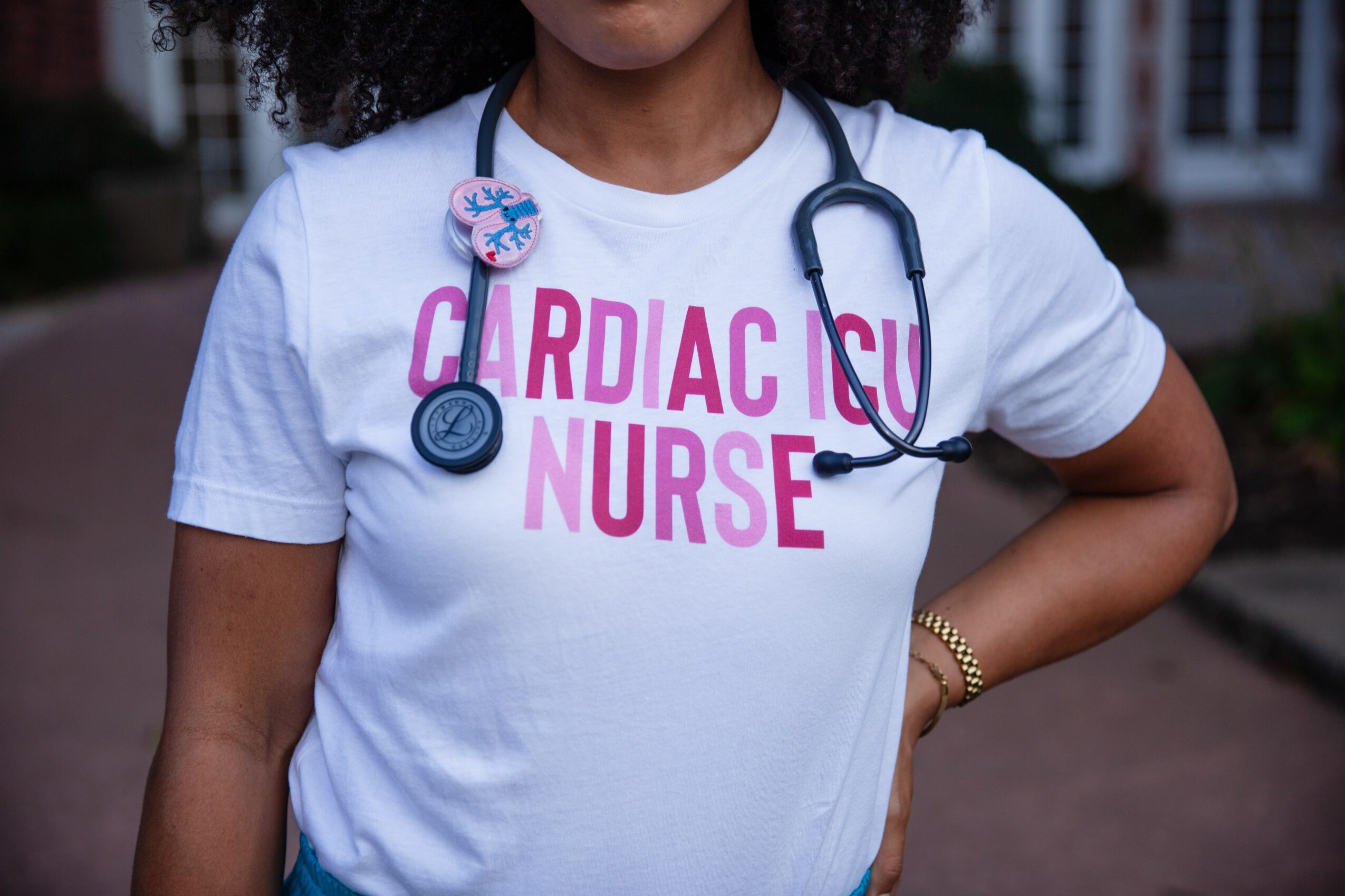 Person wearing a white T-shirt with pink text and a stethoscope around neck, standing outdoors.
