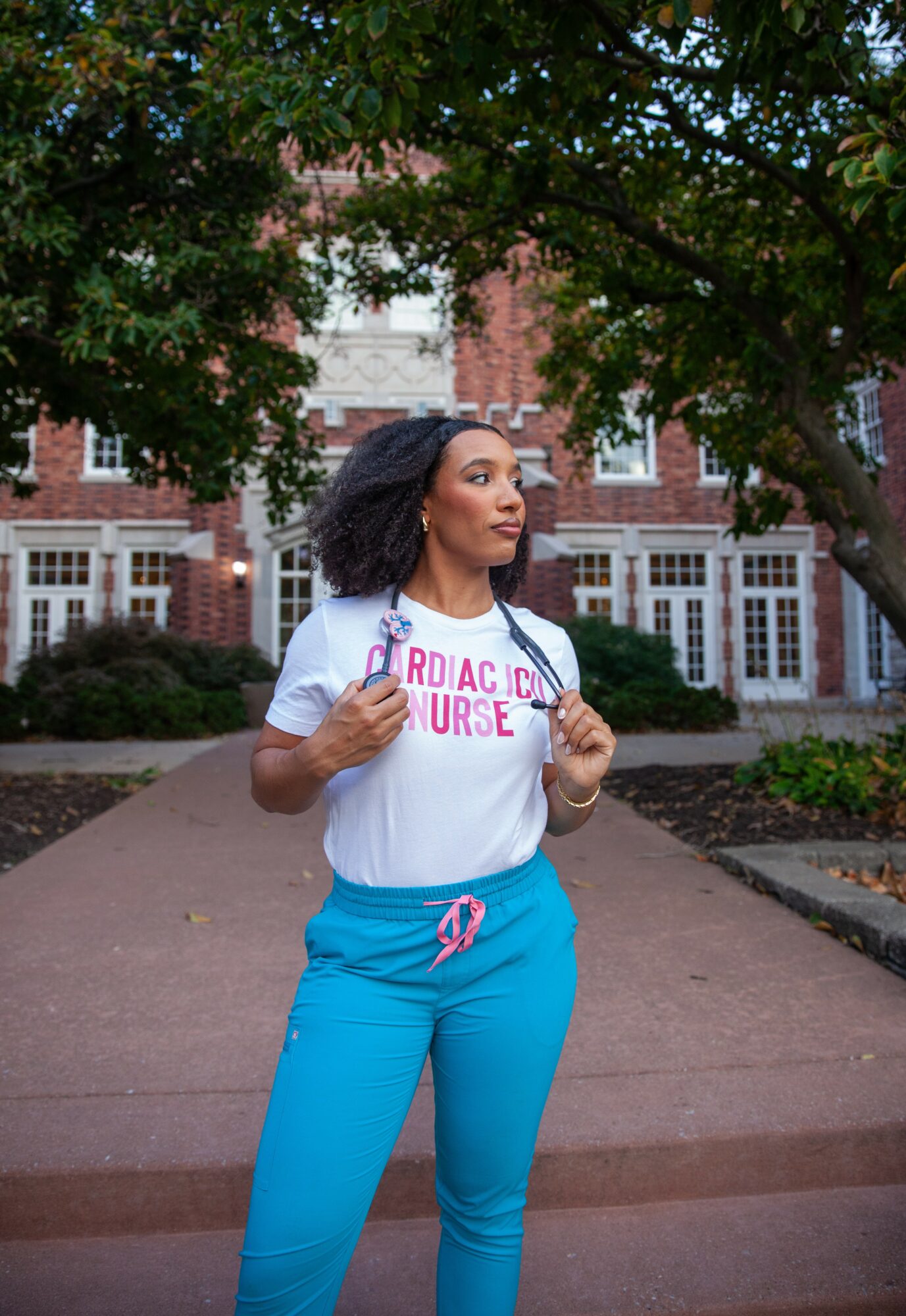 Woman standing outdoors on a pathway, wearing a white t-shirt and blue pants, with trees and brick buildings in background.