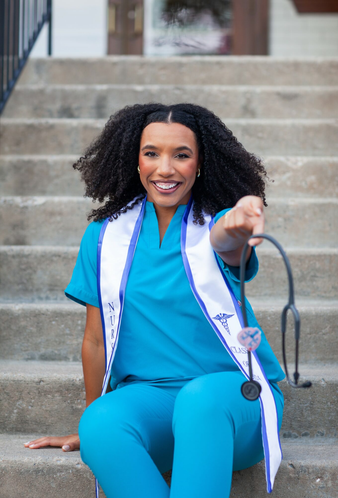 Smiling woman in blue medical scrubs sitting on stairs, pointing at the camera, with a stethoscope. Bright outdoor setting.