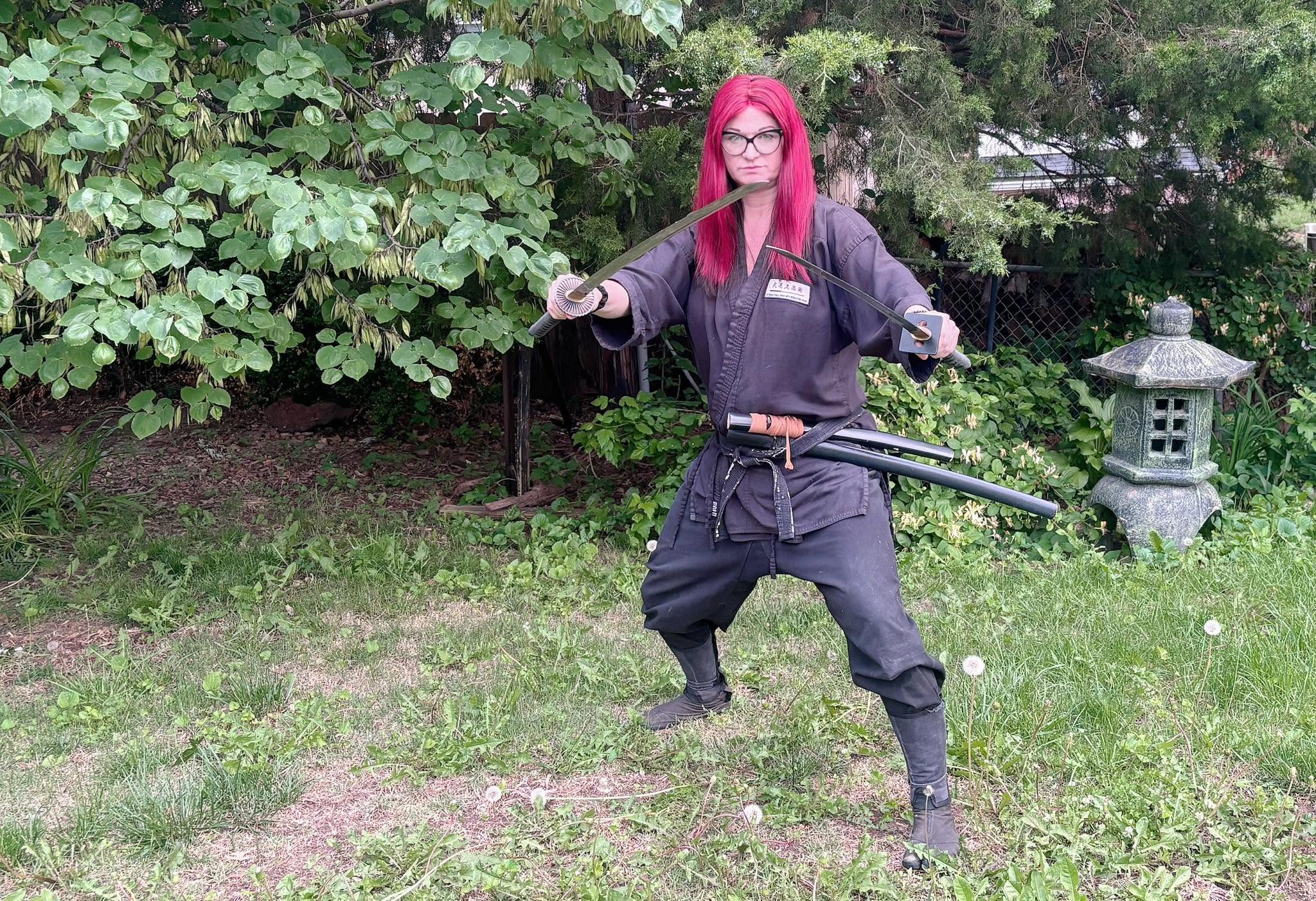 Person with red hair and glasses in a martial arts stance outdoors, holding a sword, with greenery and a stone lantern in background.