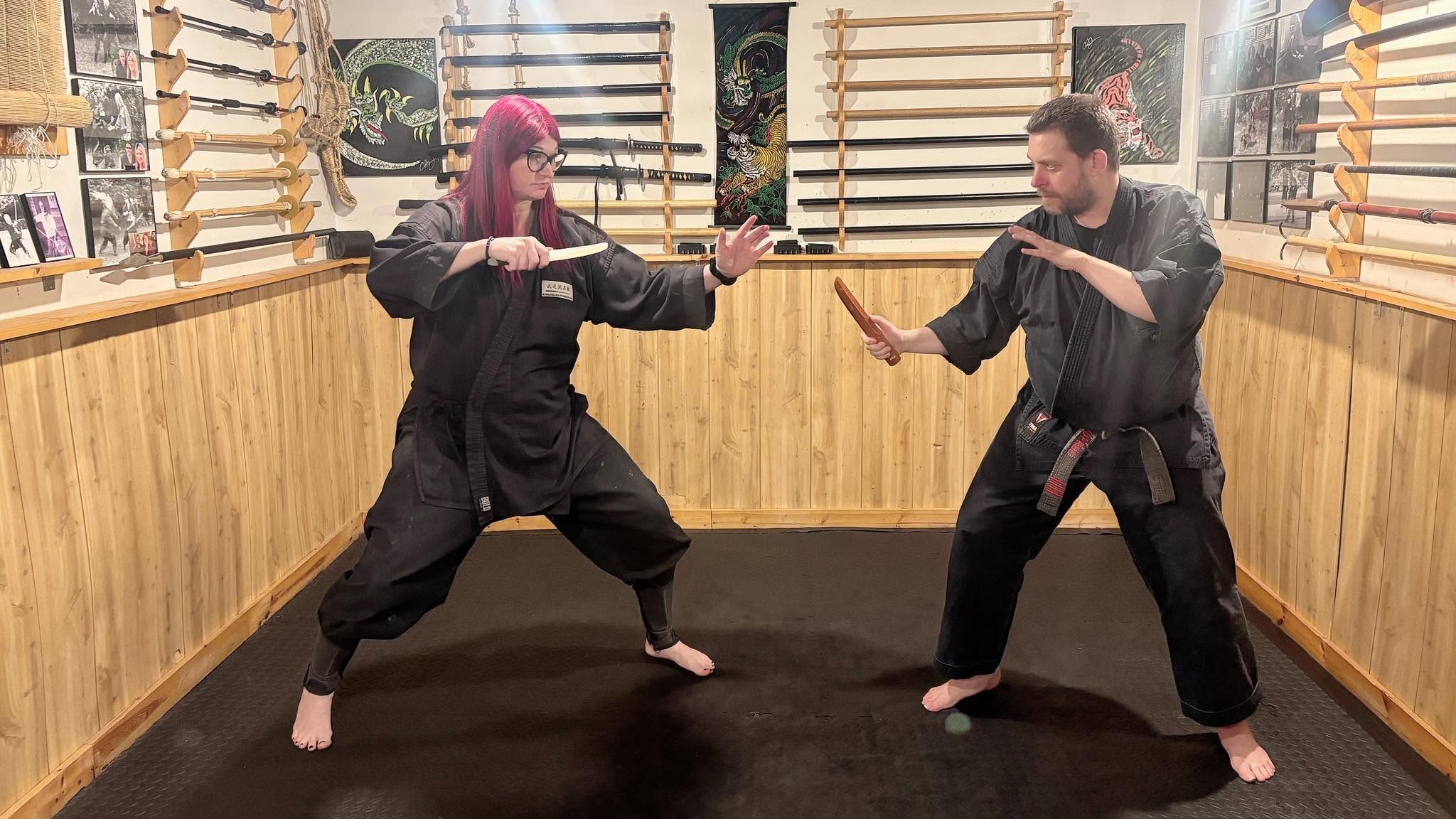 Two people practicing martial arts in a dojo, facing each other with weapons, in a room with wooden walls and shelves.
