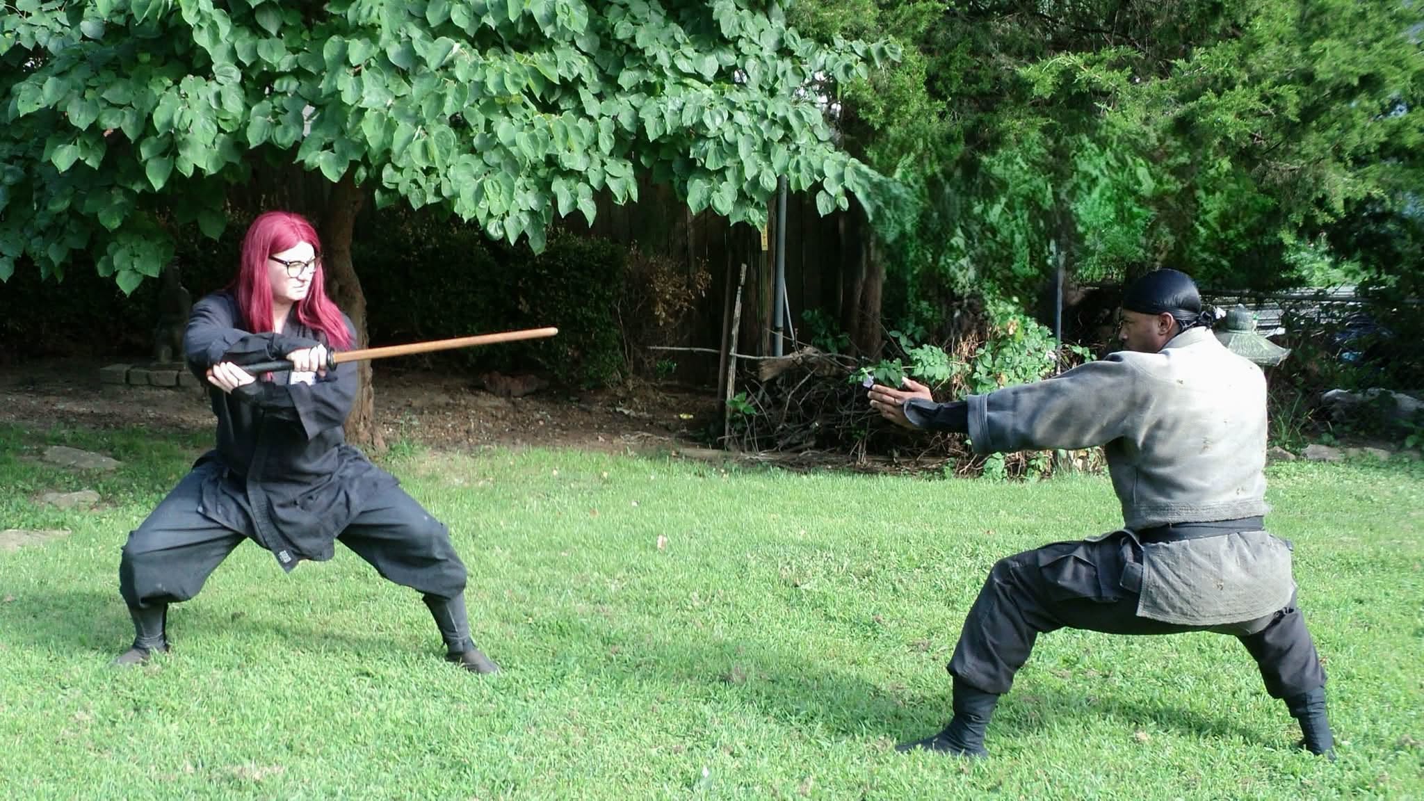 Two people in costume appear to be practicing a sword fight outdoors on grass with trees in the background.