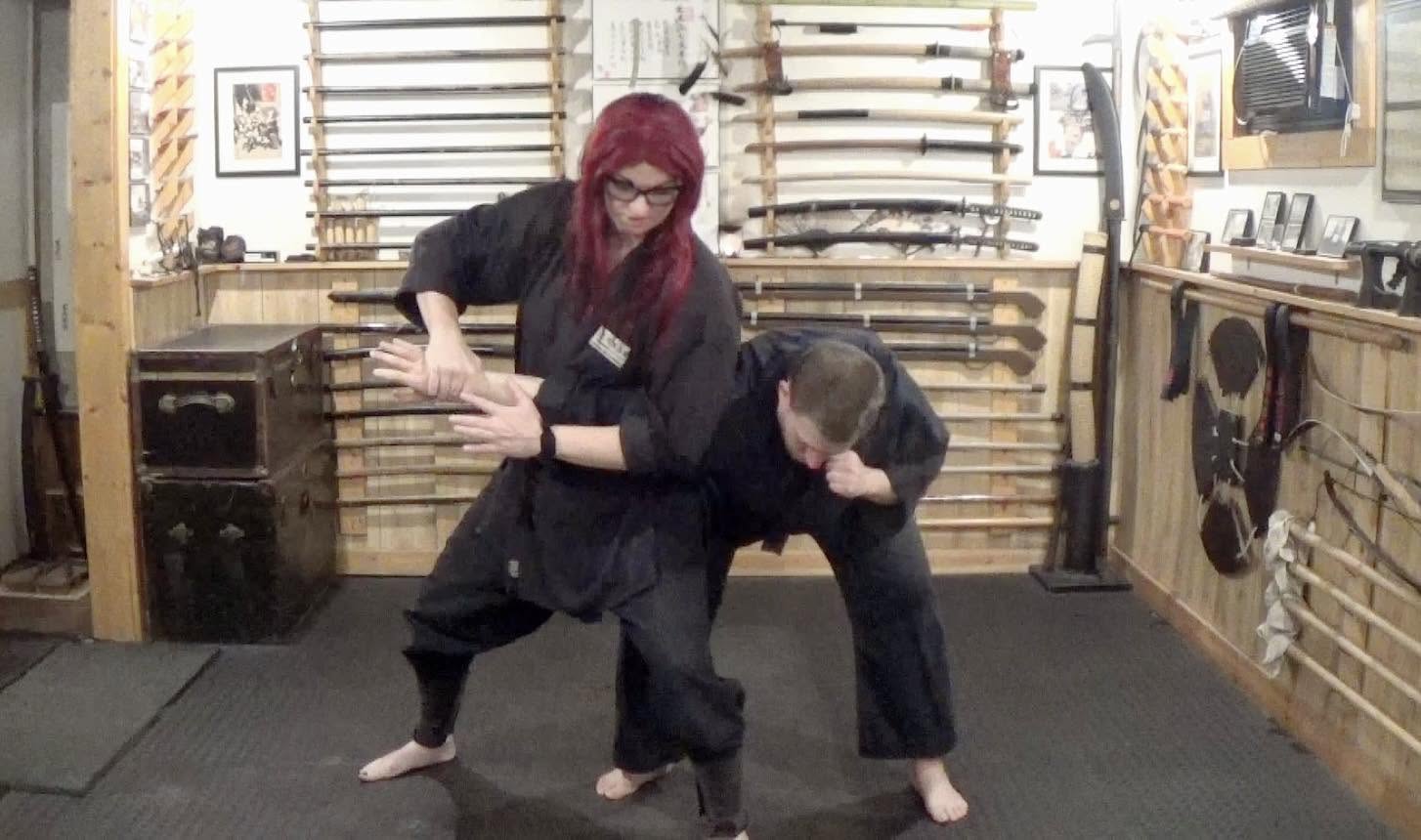 Two people practicing martial arts in a dojo, one person holding the other's arm, wooden weapons on walls.