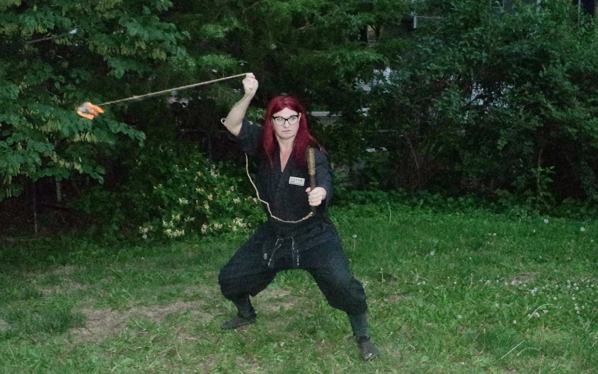 Person with red hair and glasses holding a stick in a fighting stance outdoors.