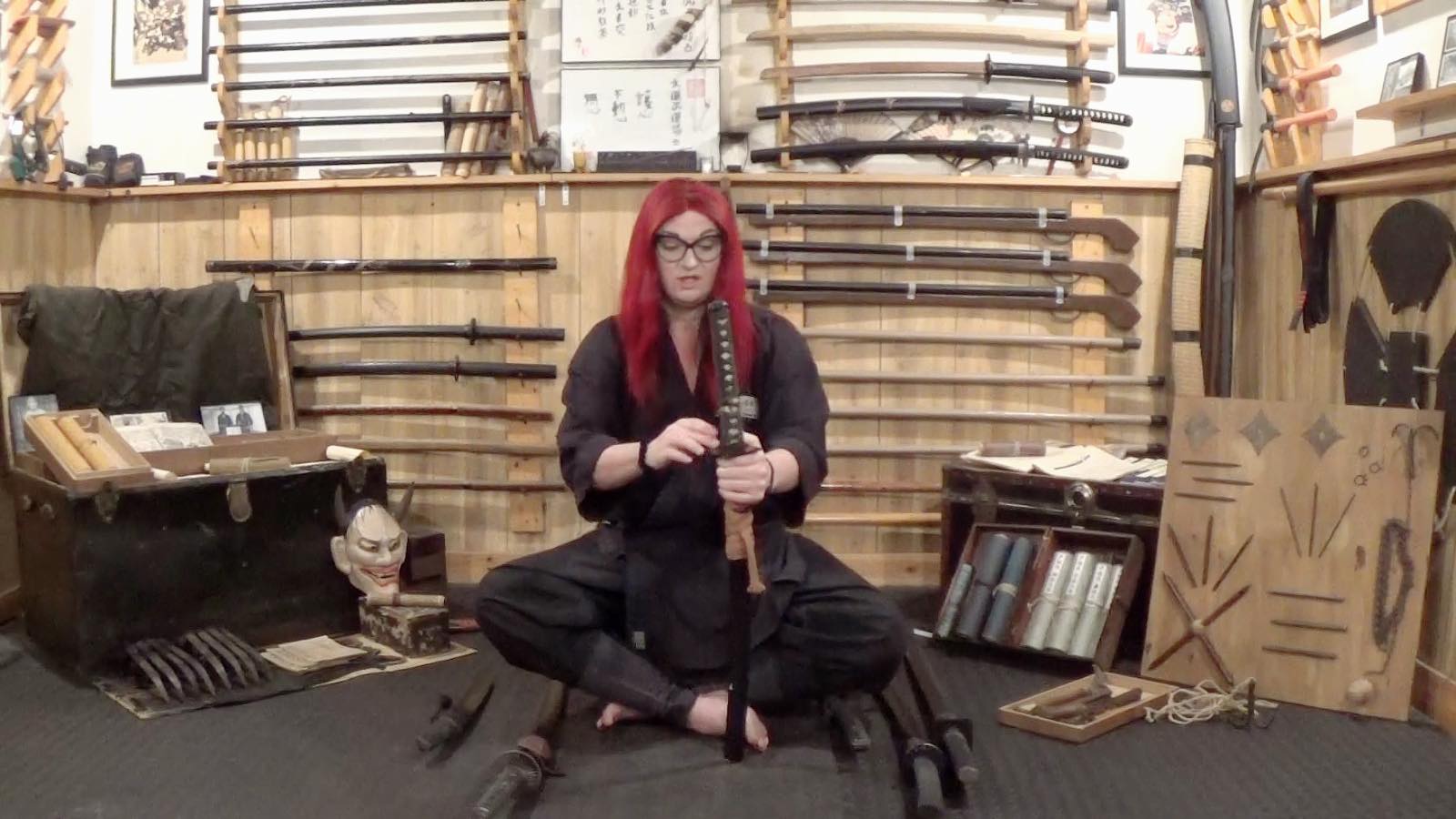 Person with red hair and glasses sitting cross-legged on the floor in a store with swords on the wall behind them.