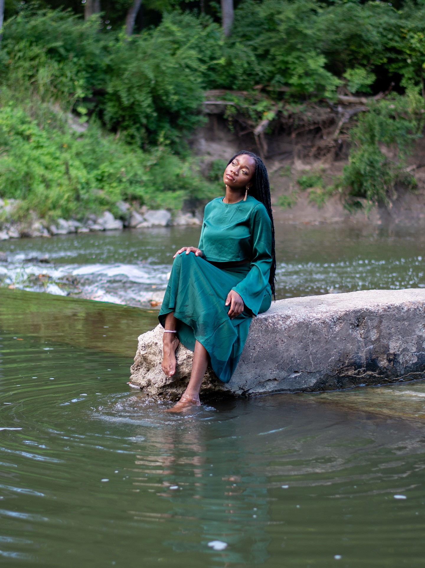 Woman in green dress sitting on a rock in a river with lush green trees in the background.