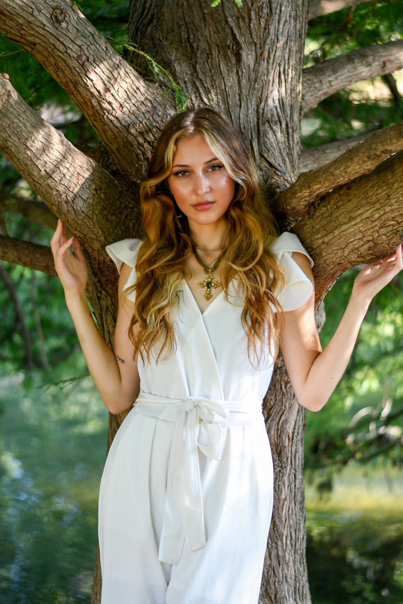 Young woman with long wavy hair in a white dress standing outdoors by a tree, with green foliage in background.
