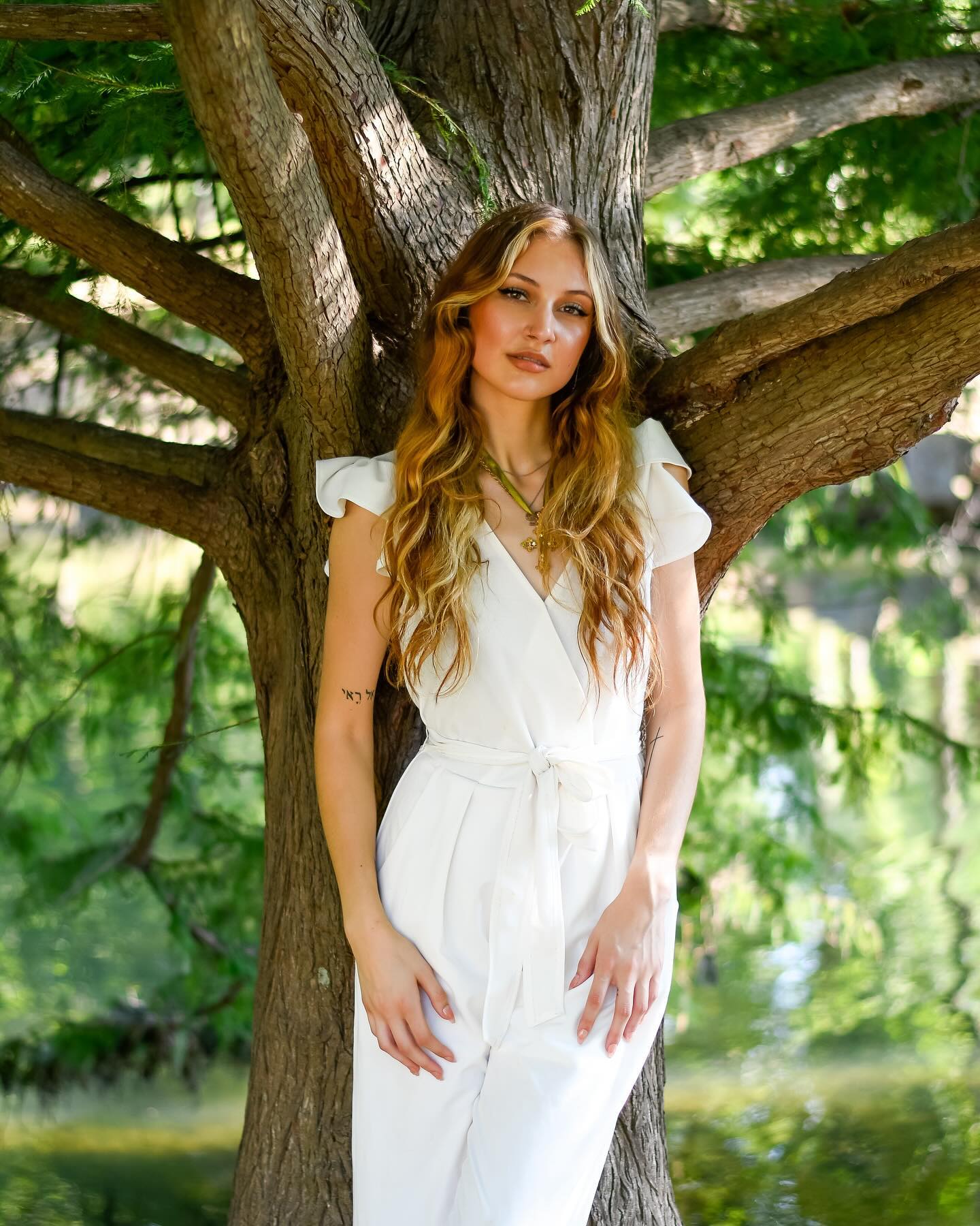 Young woman with long wavy hair in a white jumpsuit standing outdoors near a tree with green leaves.