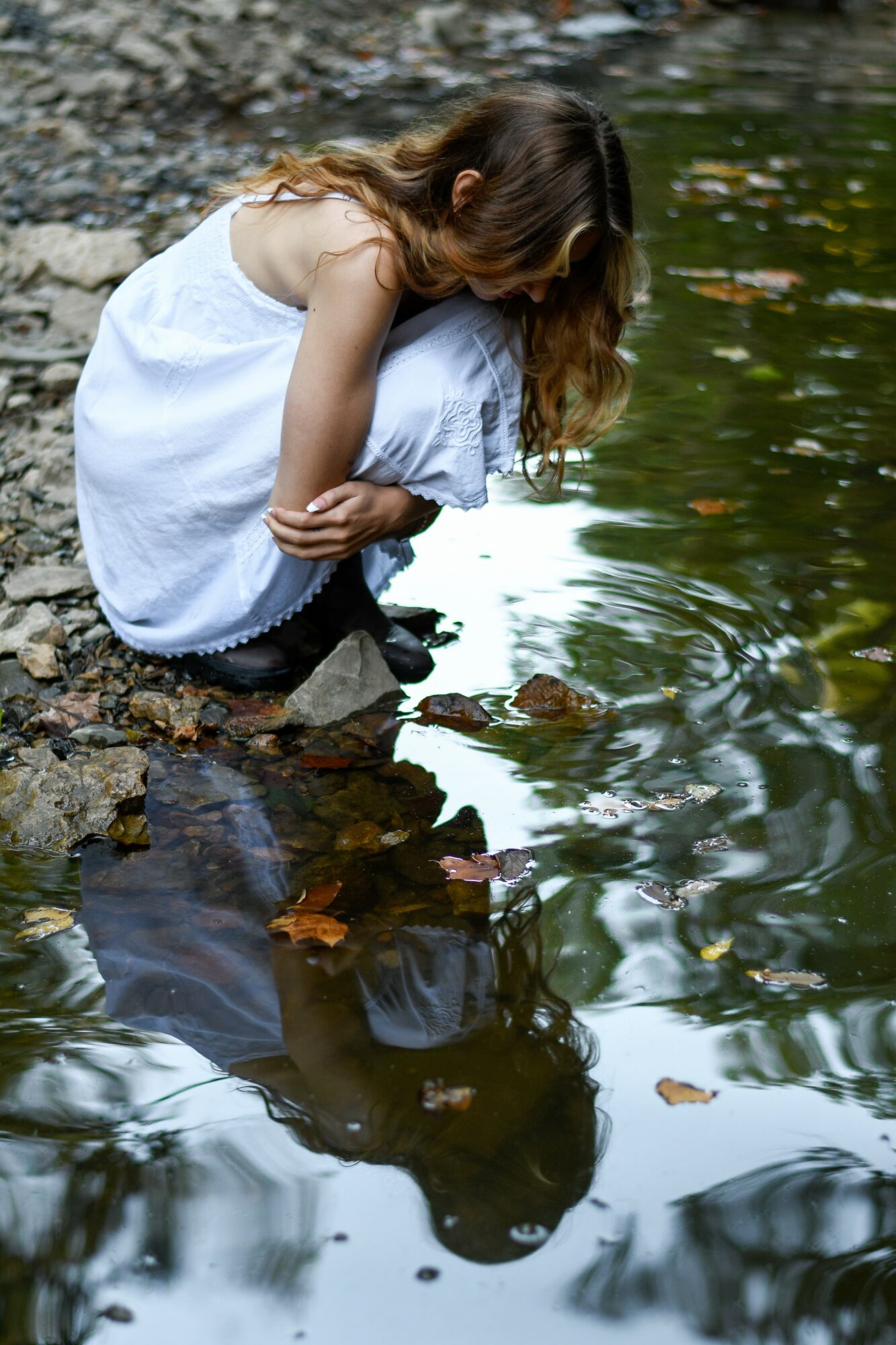 Girl in white dress crouching by water's edge, looking at her reflection, surrounded by rocks and fallen leaves.