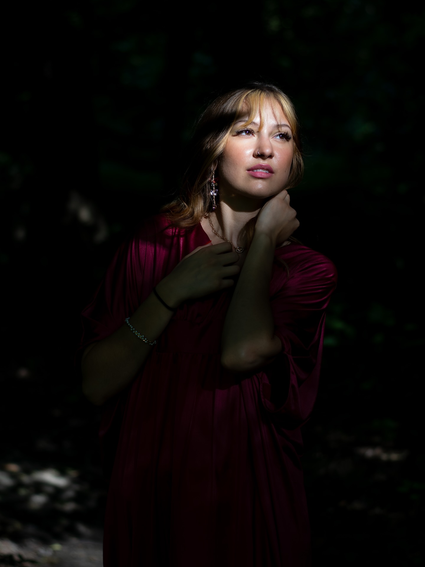 Young woman with blonde hair and earrings, wearing a red dress, standing outdoors in a dark environment, looking thoughtfully upward.