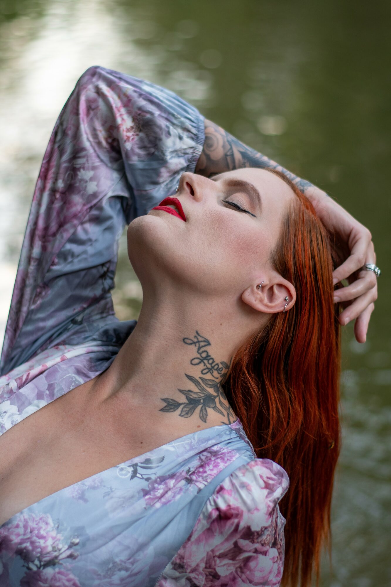 Woman with red hair and tattoo on neck, eyes closed, head tilted back, outdoors near water.