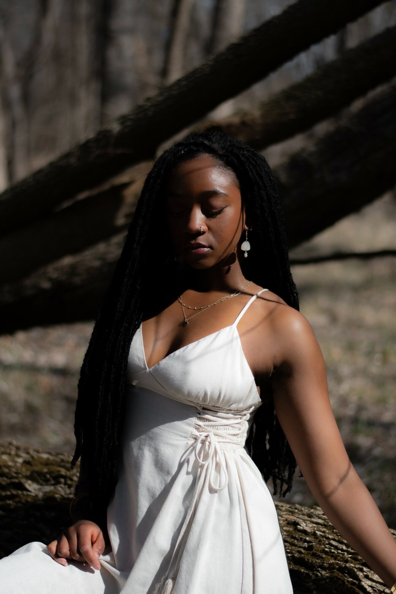 Young woman with long dark hair in a white dress outdoors, trees in background, eyes closed, serene expression.