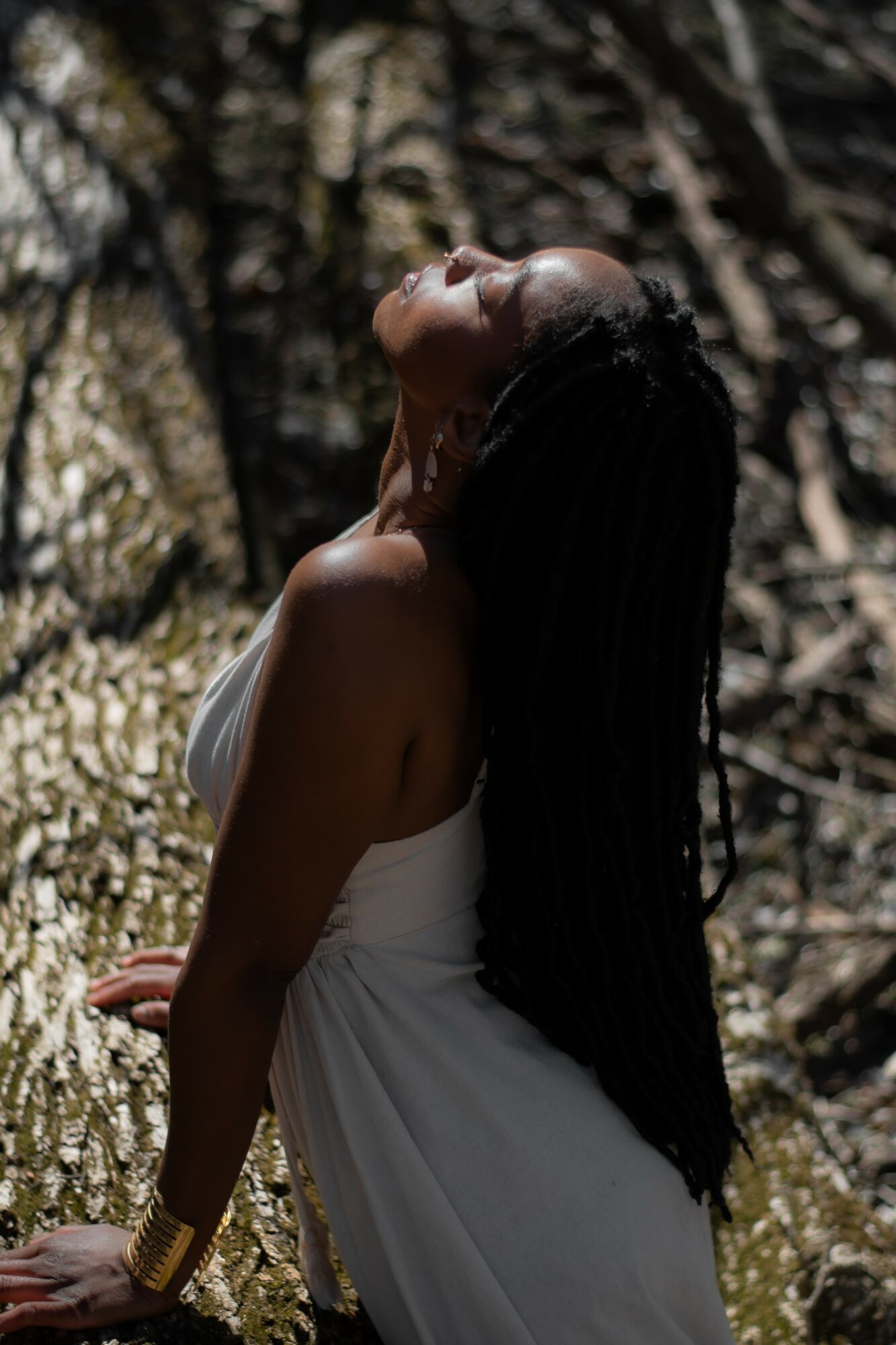 Woman with long hair tilts head back outdoors, sunlight filters through trees, wearing a white dress and gold bracelet.