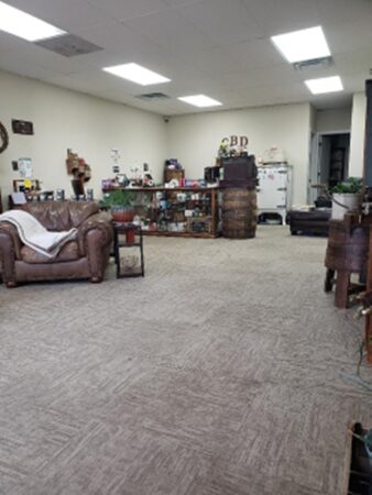 Room with a brown sofa, table, and shelves filled with various items, beige carpet, and white ceiling with lights.