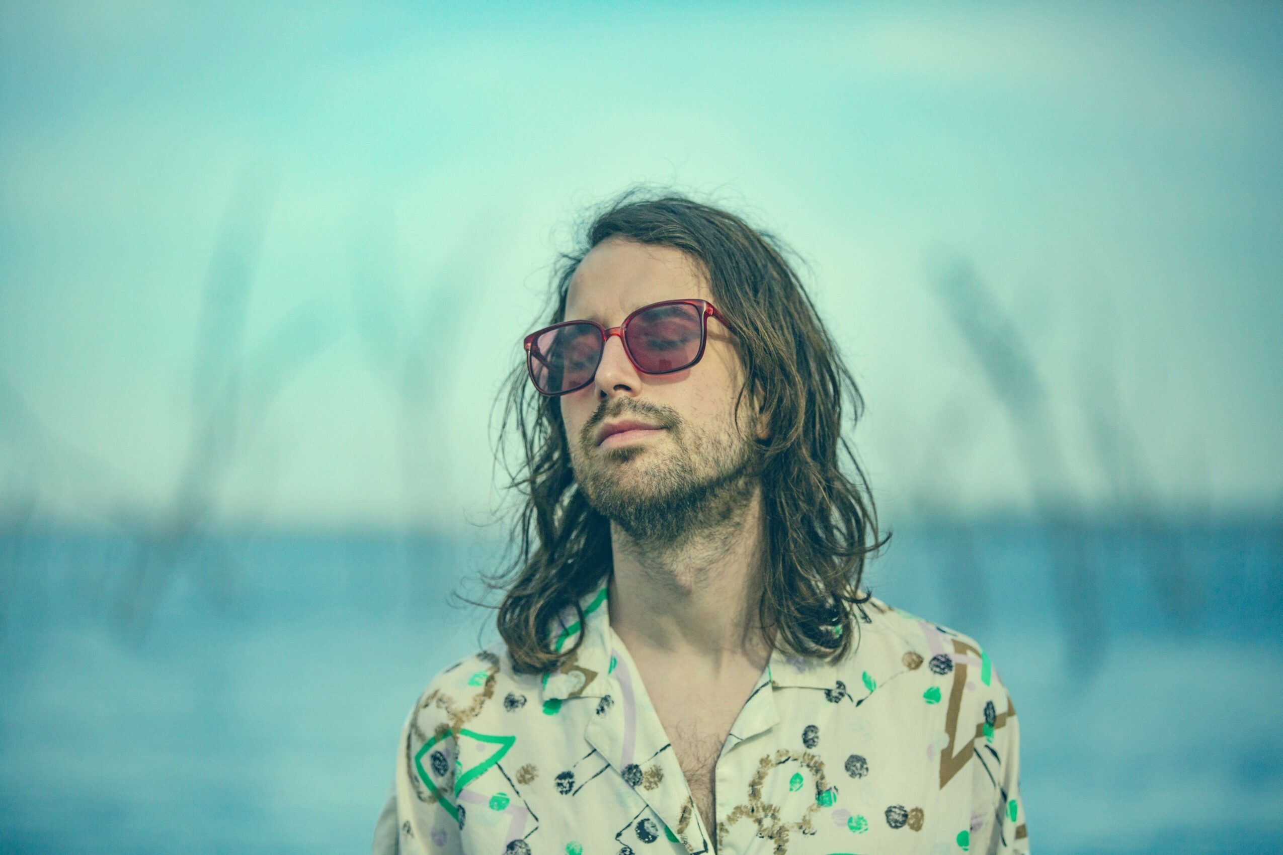 Person with long hair and sunglasses stands outdoors with a blurred water background.
