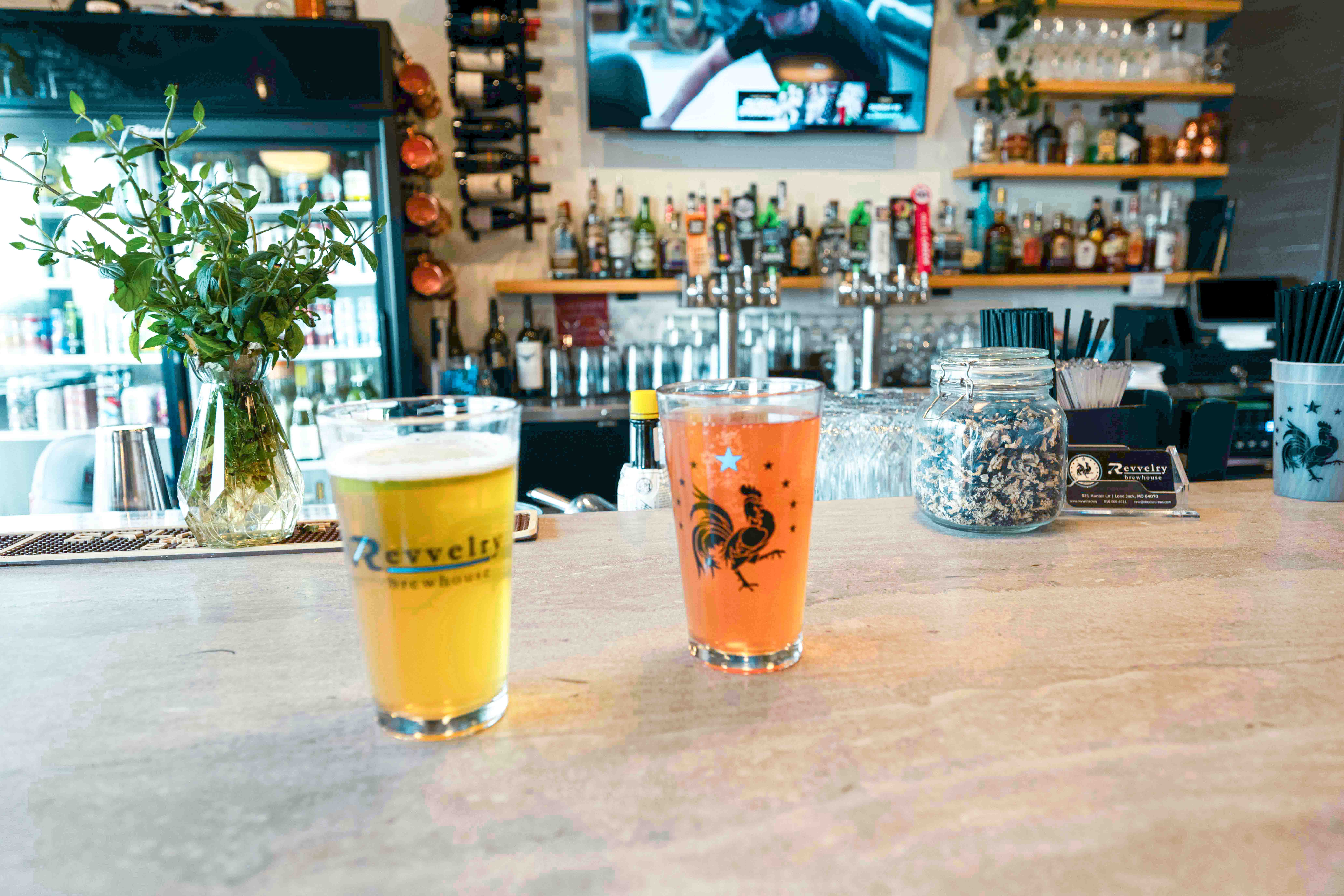 Two colorful drinks on a bar counter with a bar and TV in the background, a plant on the left, and bar supplies on the right.
