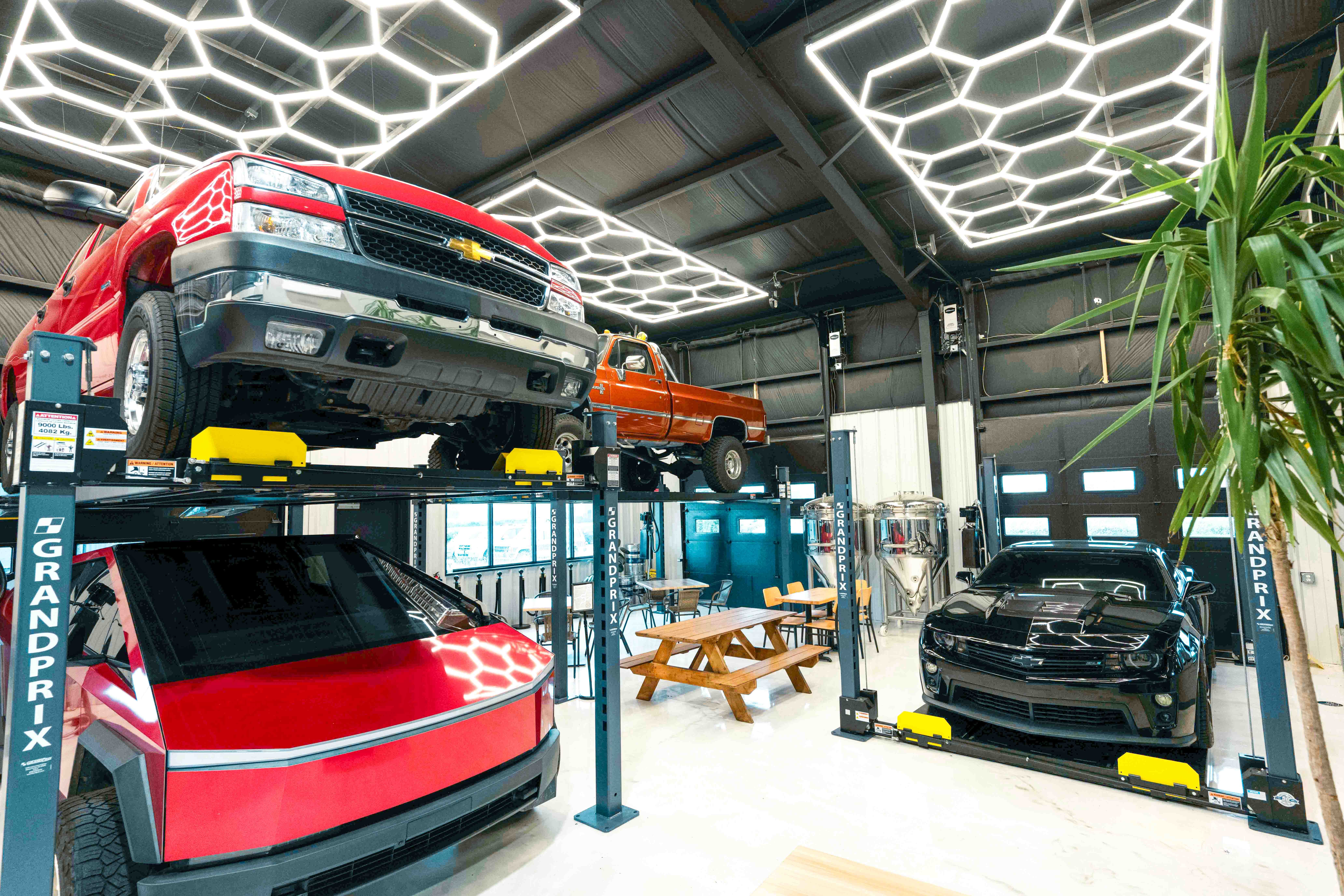 Indoor car workshop with vehicles on lifts, a picnic table, and a plant, illuminated by hexagonal ceiling lights.