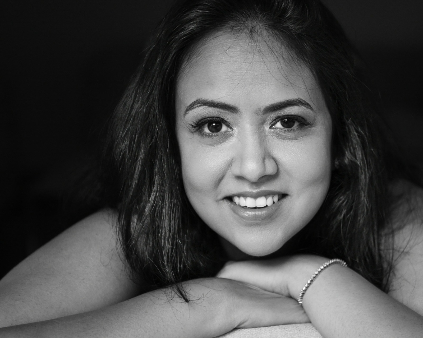 Young woman with long dark hair smiling, resting her chin on her arms, in black and white.