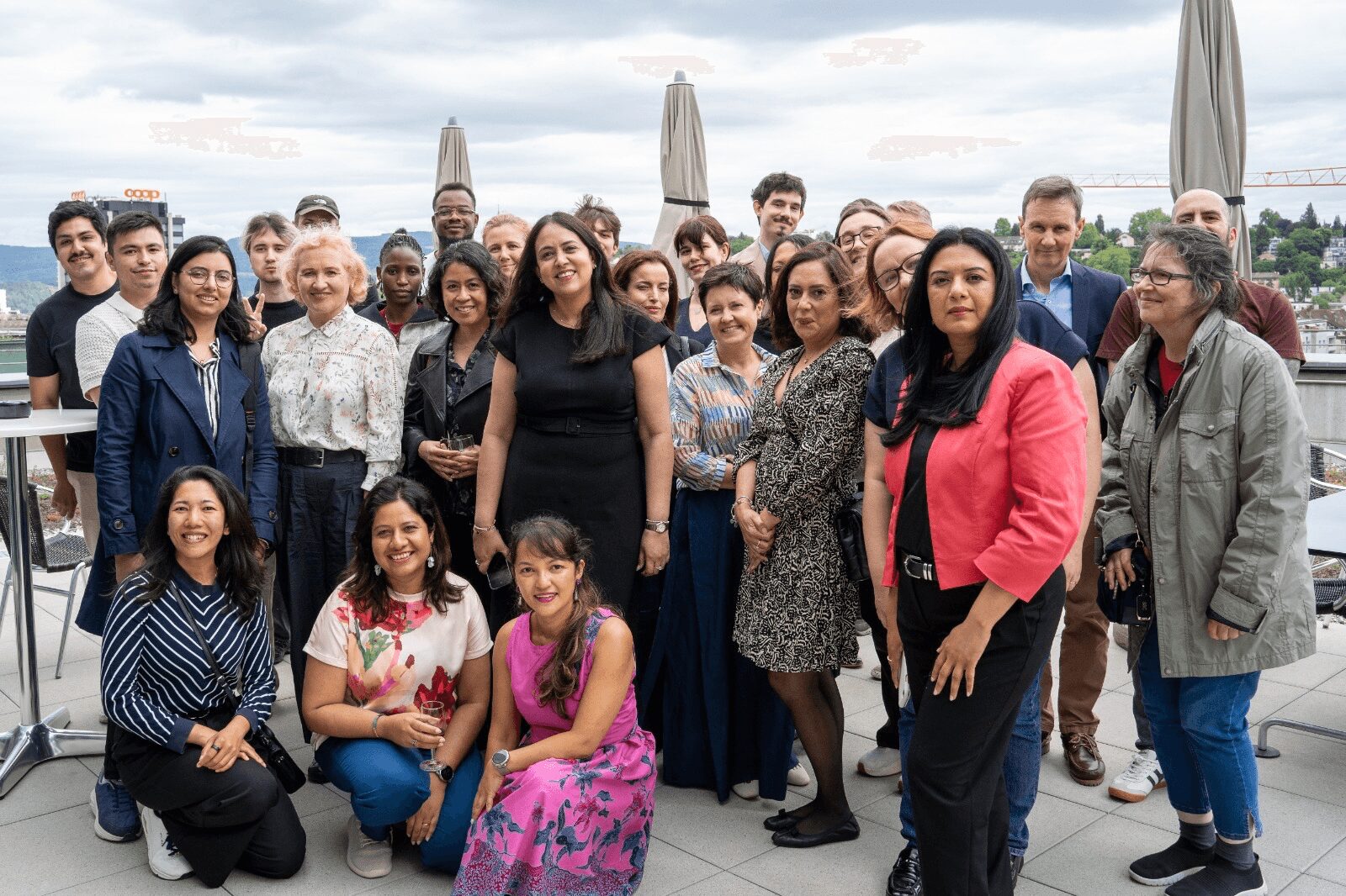 Group of diverse people standing outdoors on a rooftop with umbrellas and cityscape background.