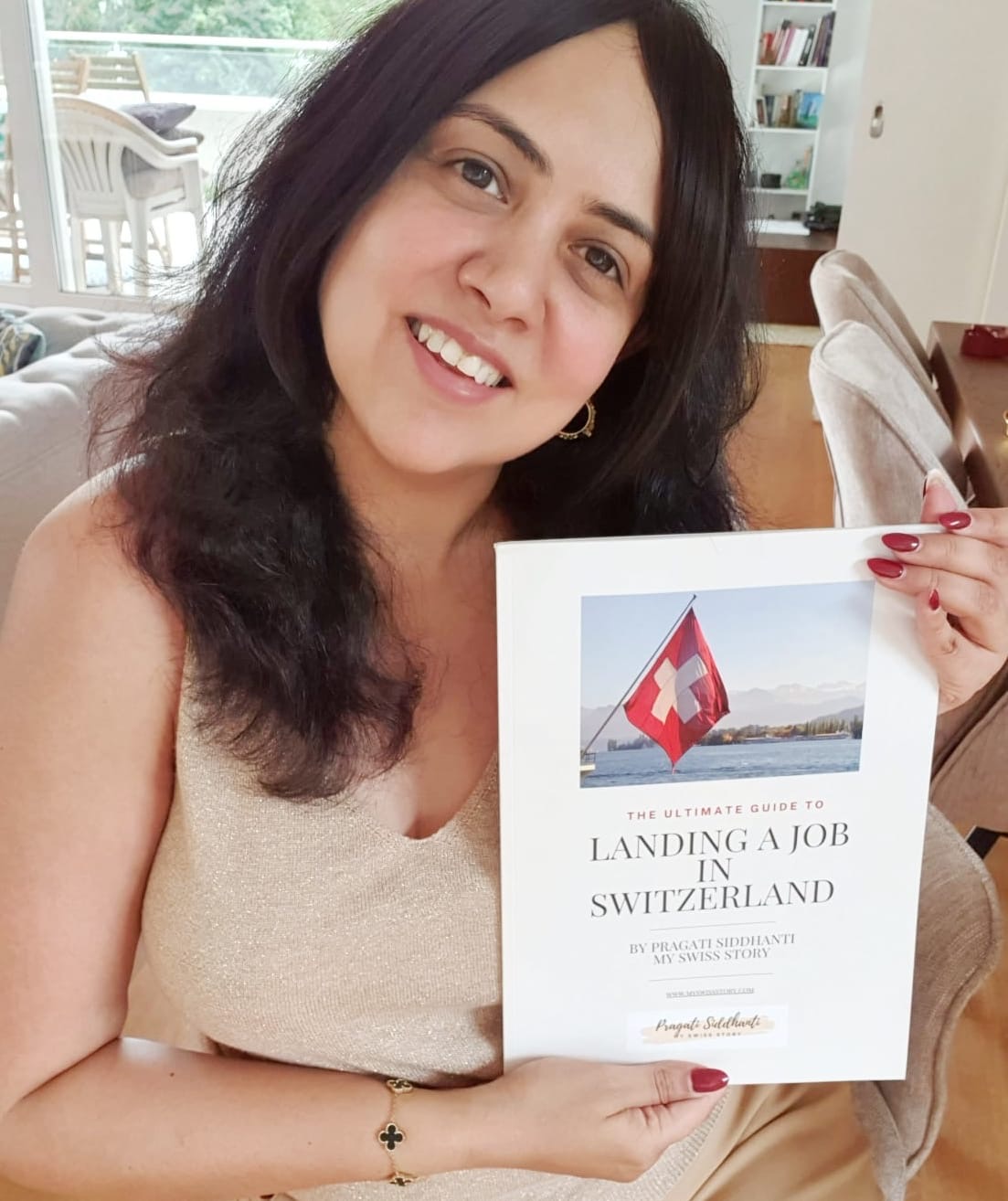 Woman with dark hair smiling, holding a book about landing a job in Switzerland, indoors with a bright background.