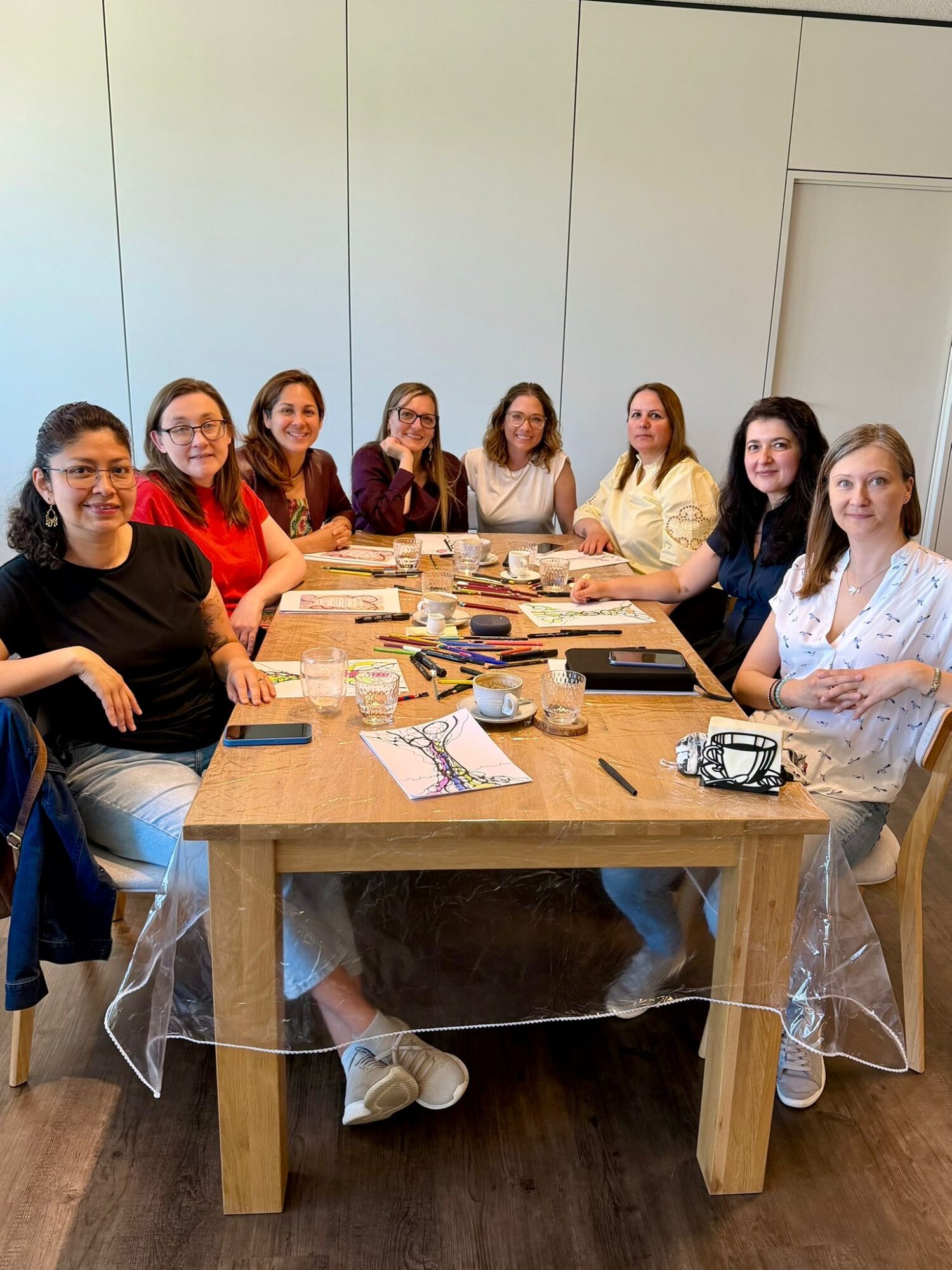 Eight women sit around a wooden table with papers, cups, and a camera, in a room with light-colored walls.