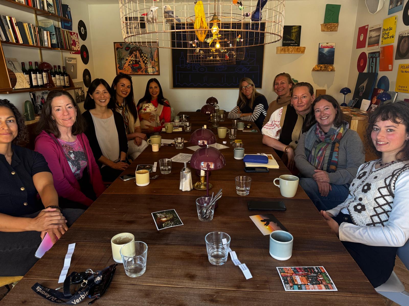 Group of people sitting around a long table in a room with colorful artwork and shelves, some with drinks and papers.