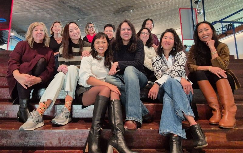 Group of ten women sitting on bleachers indoors, smiling and posing for the photo.